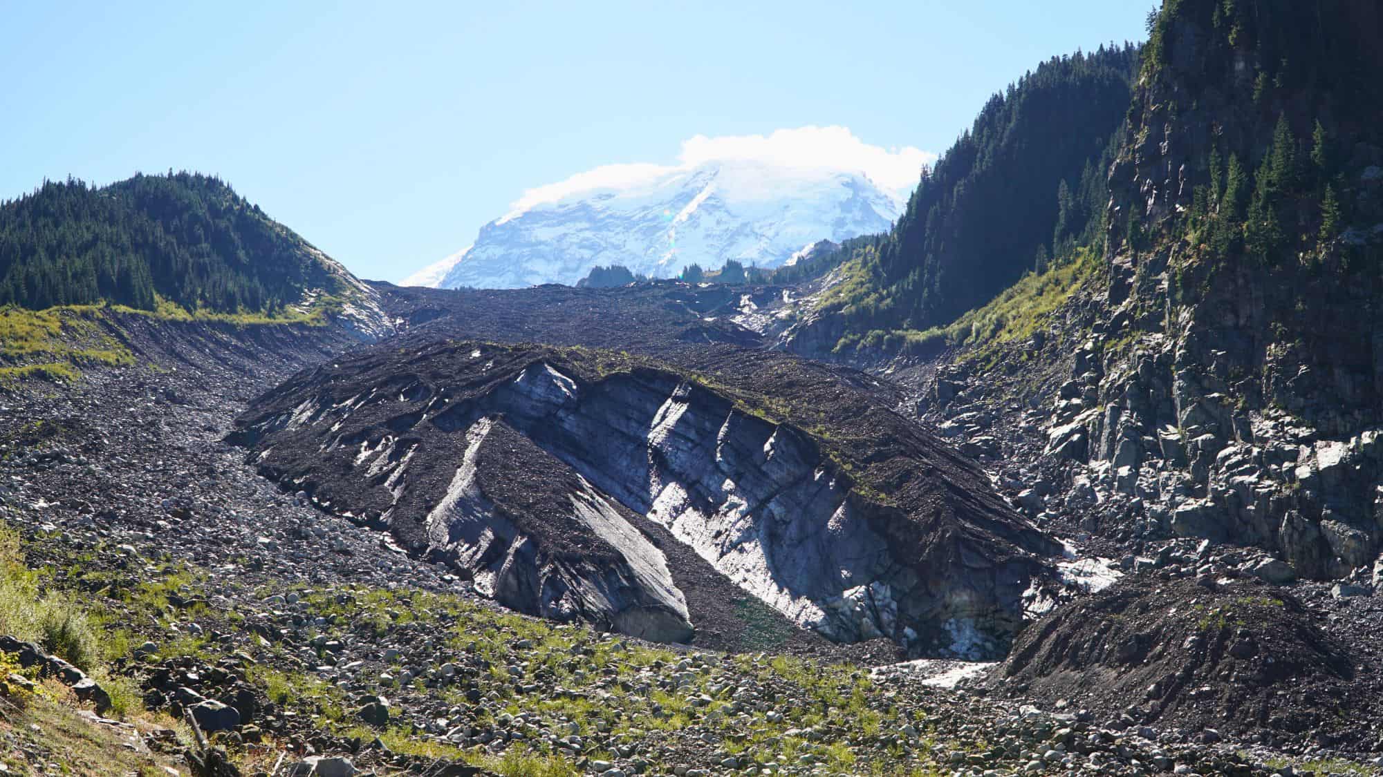 A glacier-covered mountain rises in the background behind a valley of dark rock and scree, with a ridge of ice peeking through the debris field.