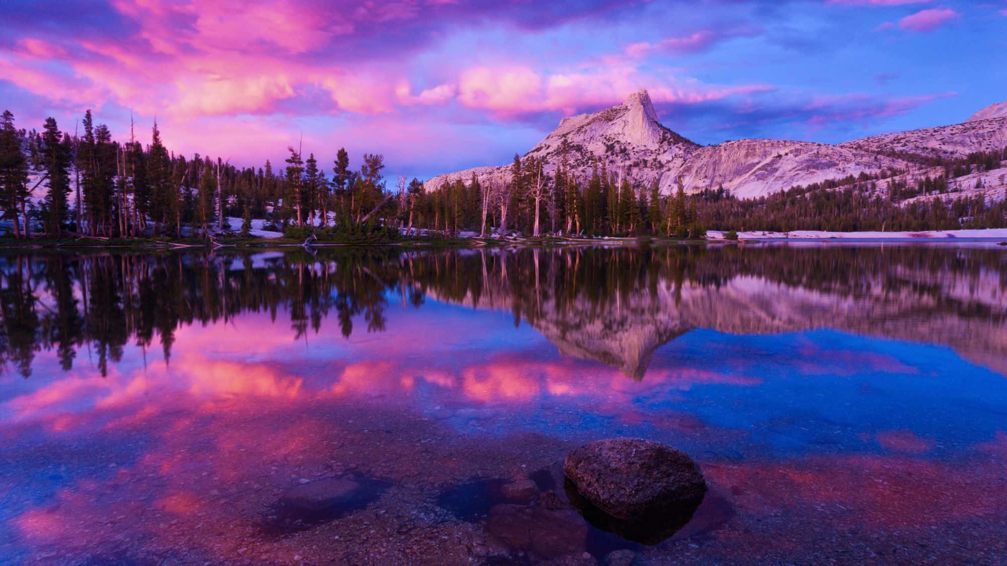 Sunset colors paint the sky in purples and pinks, reflected perfectly in the still waters of Cathedral Lake, with Cathedral Peak rising behind snow-dusted pines.