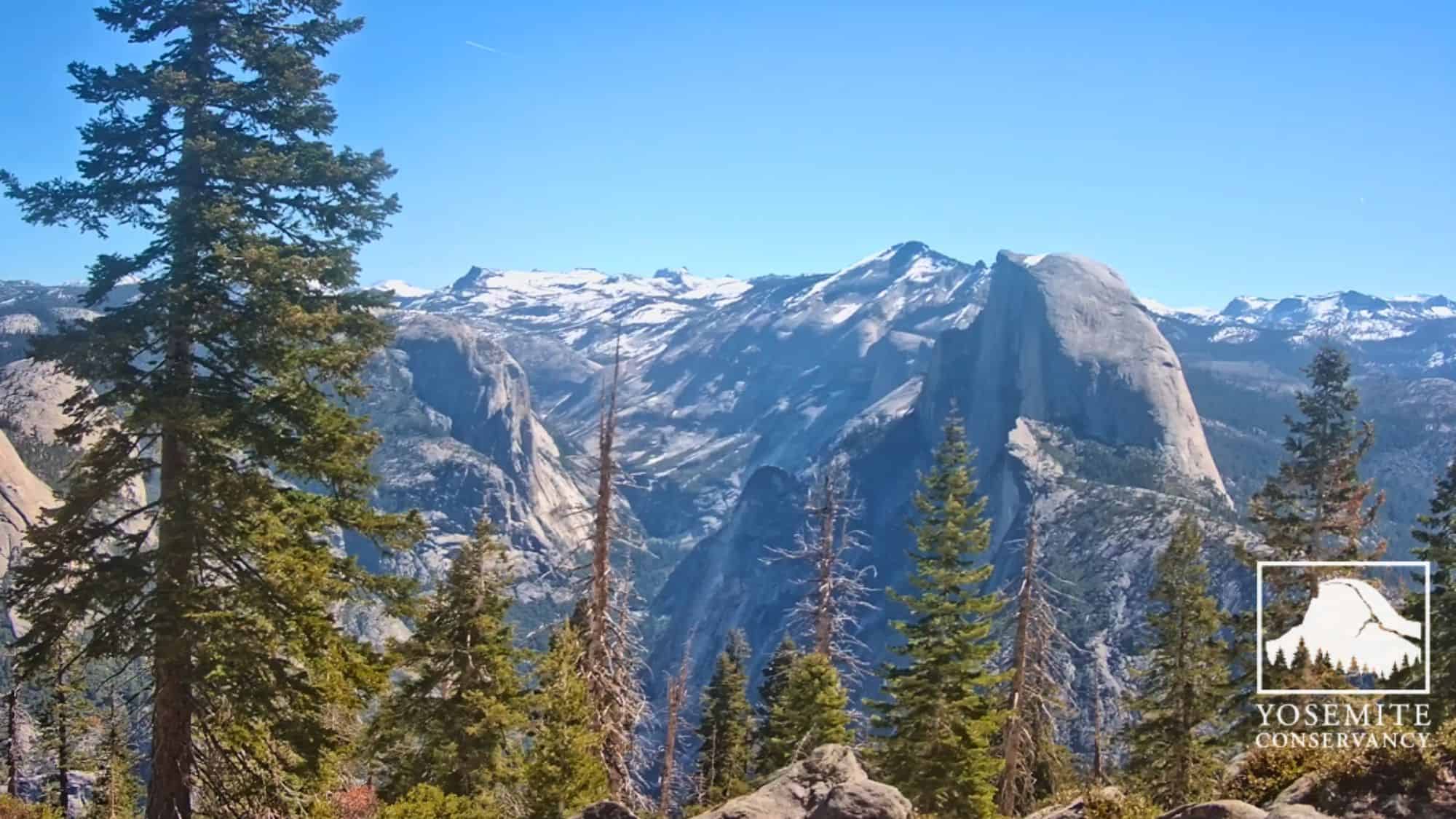 Snow-dusted Half Dome towers above the Yosemite Valley, framed by evergreen trees under a crisp blue sky, with the Yosemite Conservancy logo in the corner.