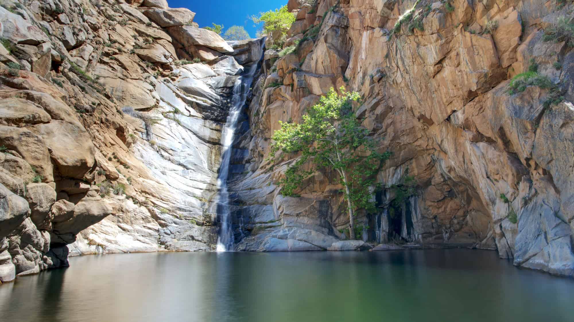 A tall, narrow waterfall cascades down rugged, tan-colored cliffs into a tranquil pool at the base, with sparse greenery and a bright blue sky overhead.