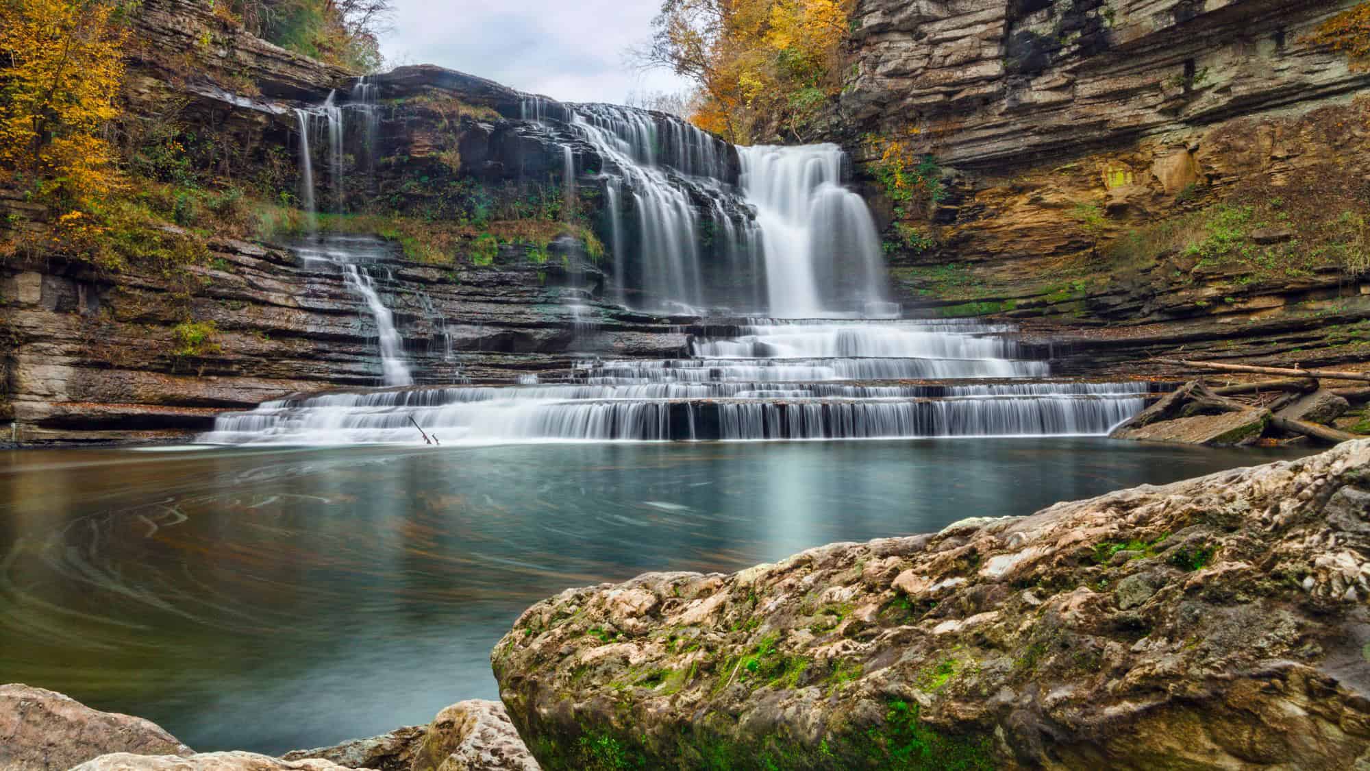 A gorgeous multi-tiered waterfall cascading over rock ledges into a large plunge pool, surrounded by autumn-colored trees and rugged cliffs.