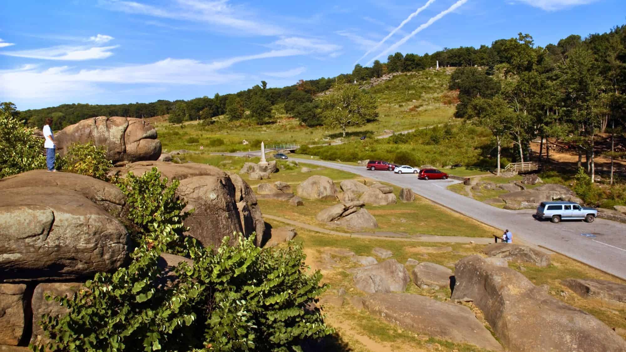 Scattered boulders and a few visitors overlook a battlefield from the famous Devil’s Den rock formation at Gettysburg, with cars and monuments visible in the background.
