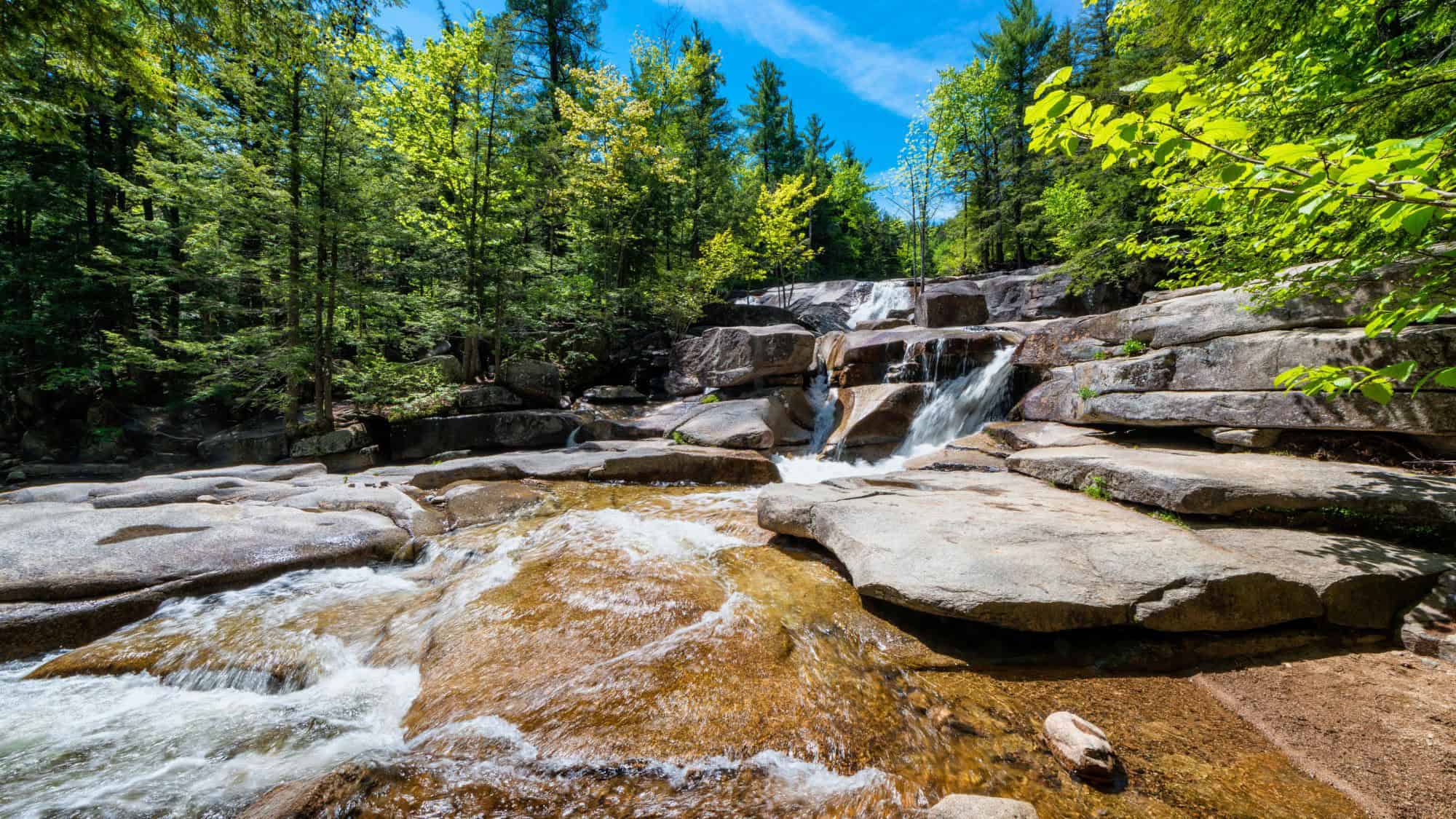 A series of small, cascading waterfalls tumbling over granite rocks in a sun-dappled forest, creating shallow pools.