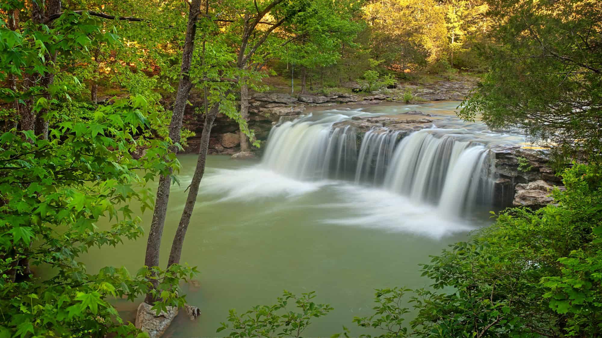 A picturesque waterfall flowing over a rock shelf into a serene green pool, framed by vibrant spring foliage.