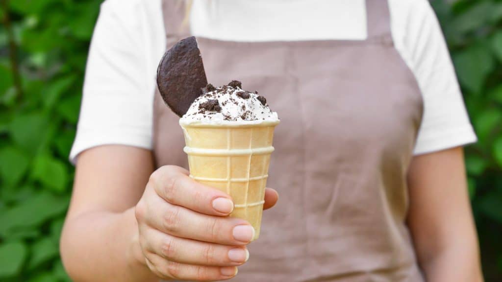 A hand holds out a waffle cone topped with cookies and cream ice cream, garnished with crumbled cookie bits and a whole chocolate cookie against a leafy green backdrop.