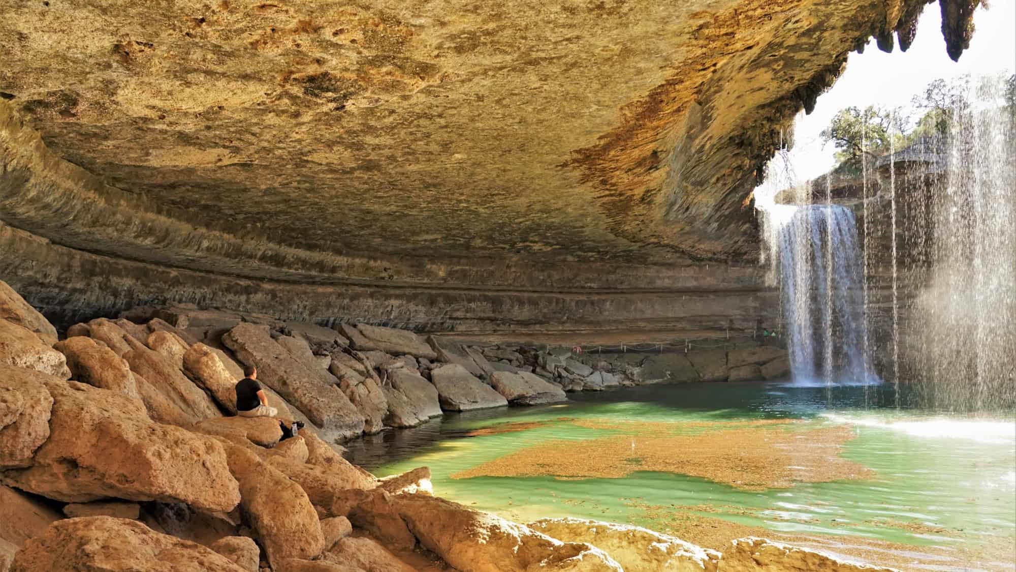 A stunning grotto with a natural pool beneath a massive rock overhang, where a thin waterfall spills into emerald-green water surrounded by boulders.