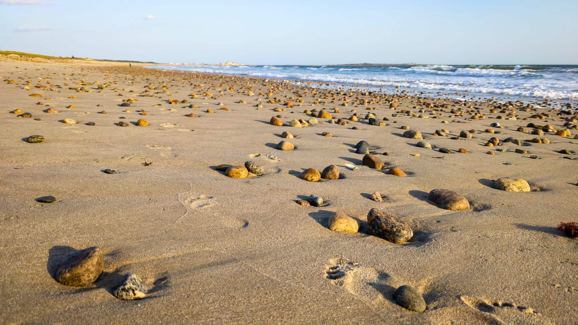 Close-up of a sandy beach covered in scattered round stones of varying colors, with soft waves lapping in the background and footprints in the sand.