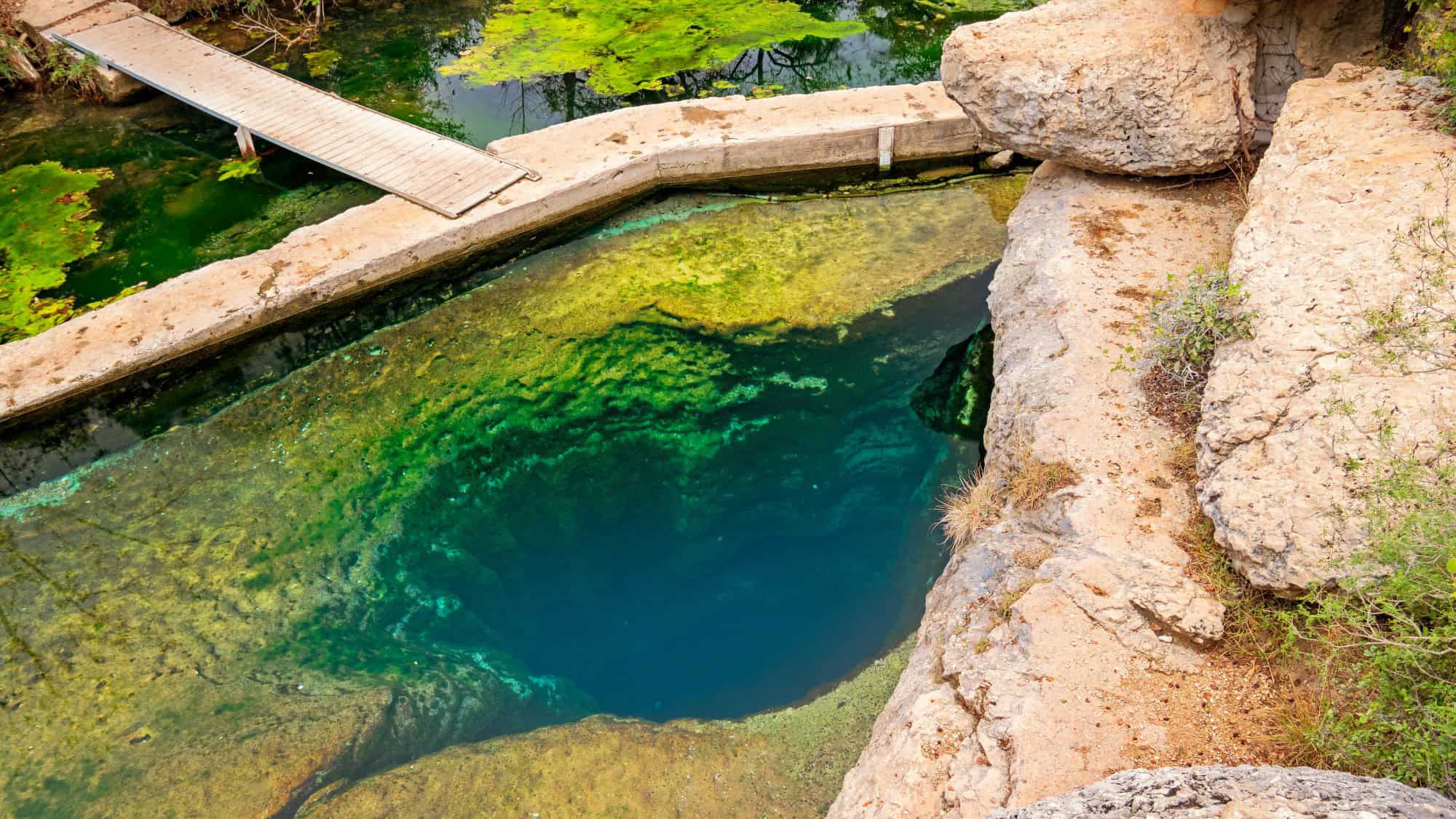 A striking natural artesian spring with a deep vertical shaft, surrounded by limestone rocks and clear green-blue water.