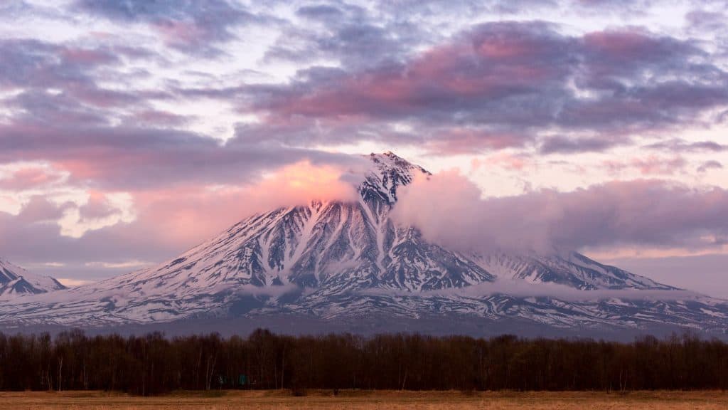 Koryaksky Volcano towers above the Kamchatka Peninsula, its snowy ridges catching the soft pink light of sunset, with scattered clouds enhancing the dramatic scene.