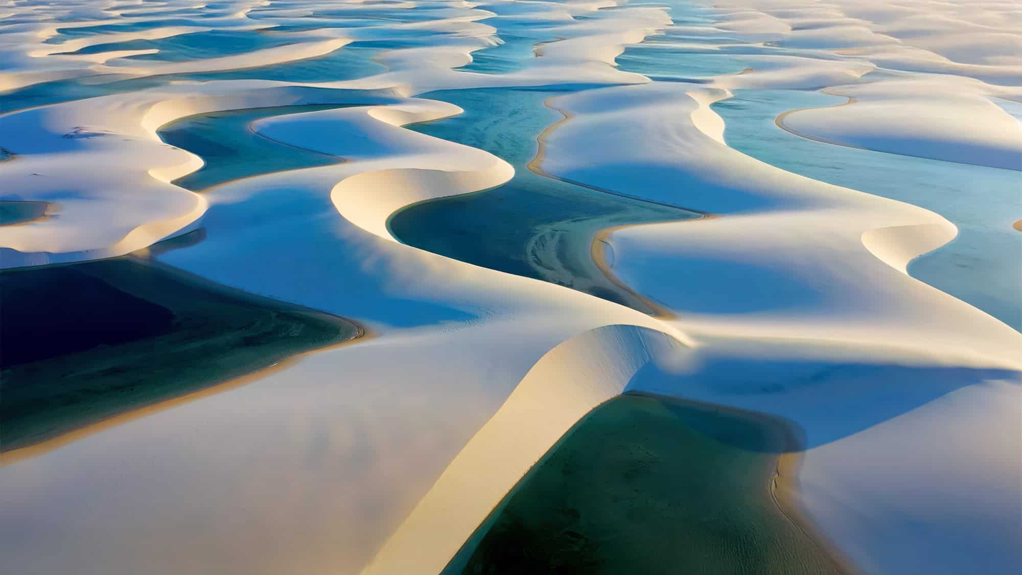 An aerial view of Brazil's Lençóis Maranhenses National Park shows sweeping white sand dunes filled with winding, crystal-blue freshwater lagoons, creating a surreal landscape of rippling curves and contrast.