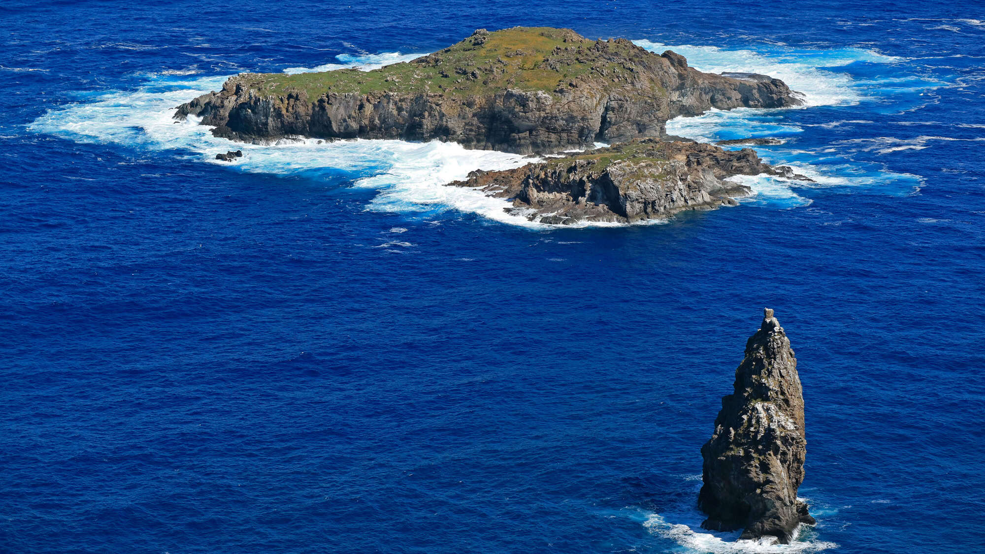 A rugged, remote view of Motu Nui and its surrounding rocky islets, surrounded by vibrant blue ocean waves crashing against the cliffs.