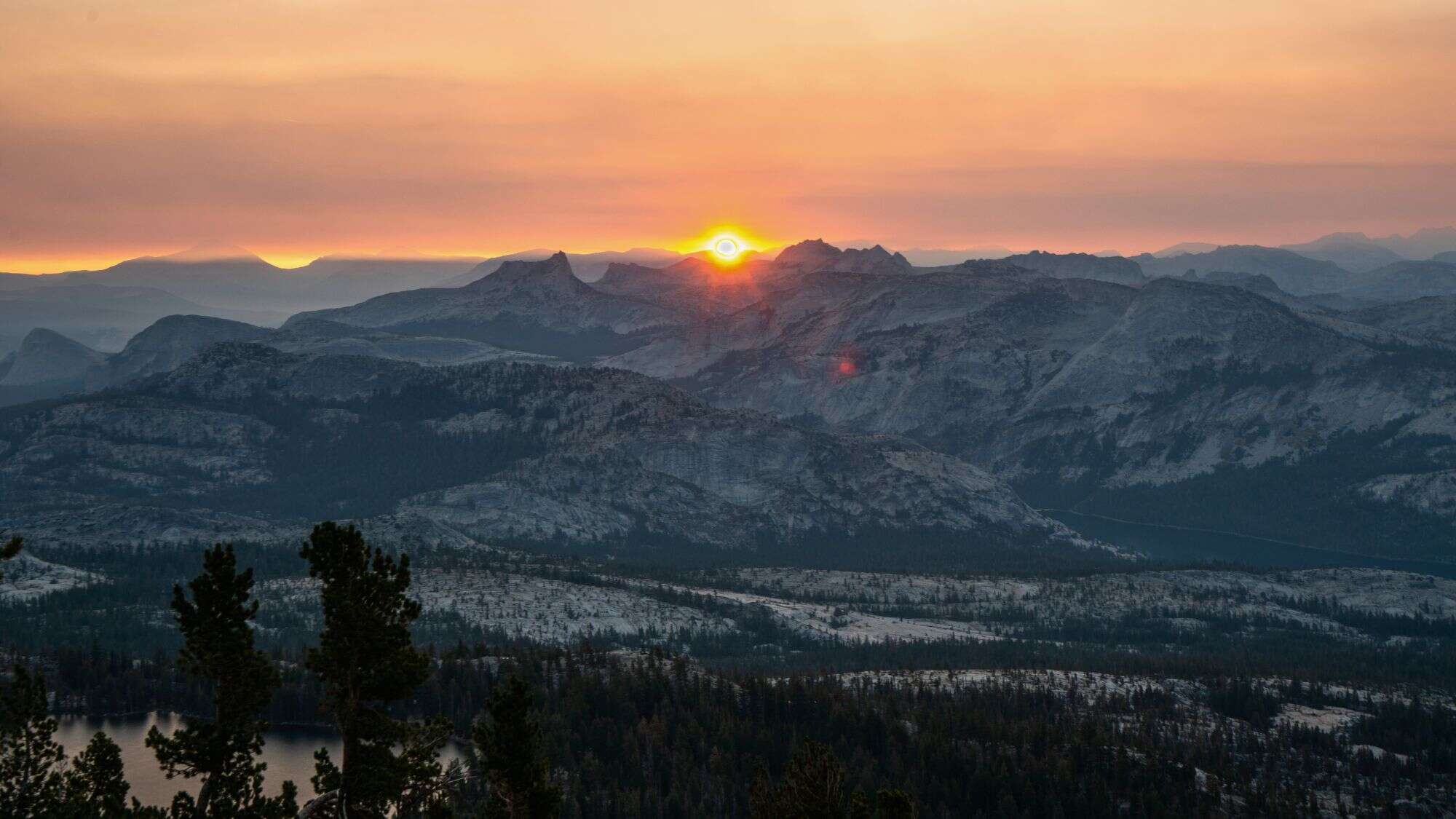 A glowing sunset dips below the horizon, casting warm orange hues across the jagged ridgelines of Mt. Hoffman and the surrounding peaks in Yosemite.