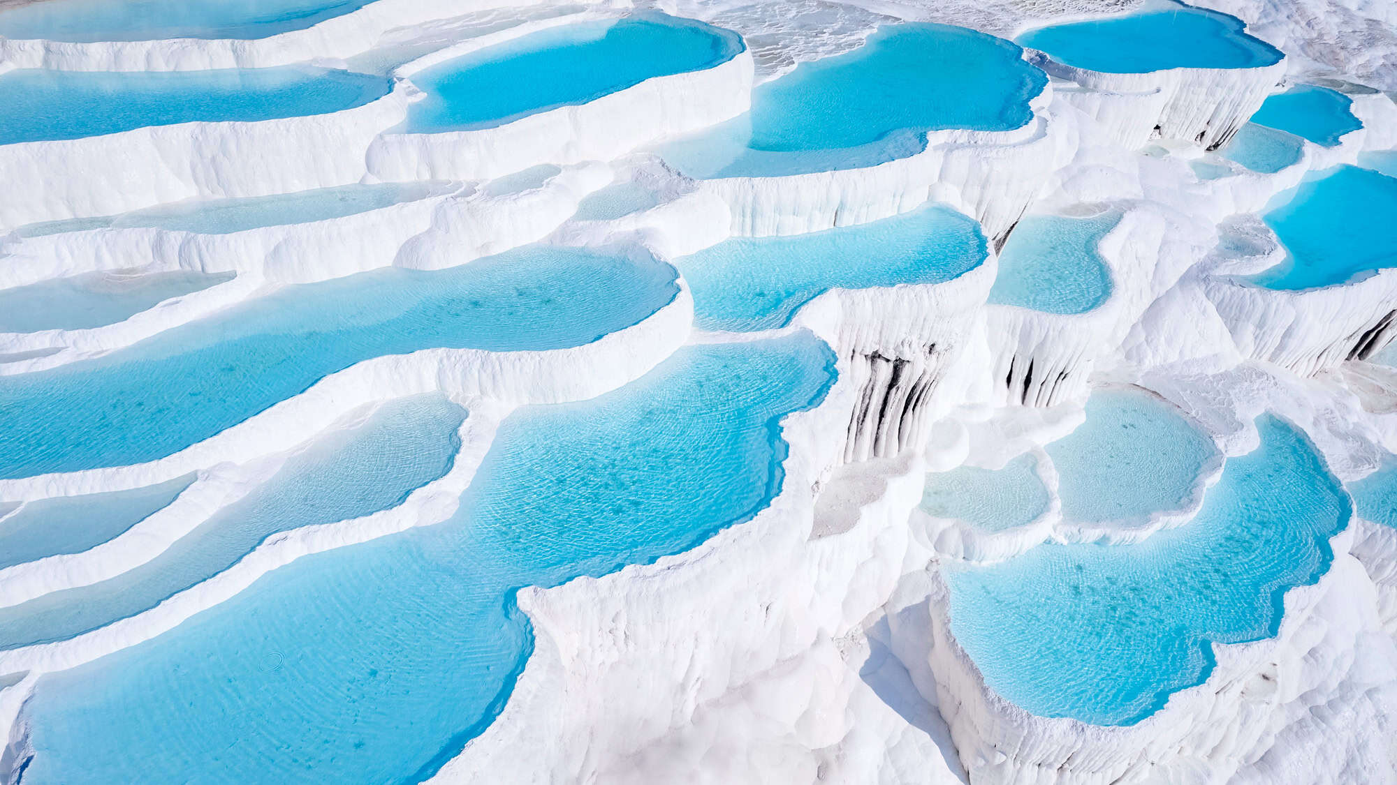 Aerial view of Turkey's Pamukkale reveals a dazzling staircase of white travertine terraces filled with bright turquoise thermal waters, shaped by centuries of mineral-rich hot springs.