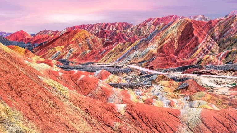 A vibrantly colored mountain range showcases exaggerated hues of red, orange, yellow, and lavender under a soft pink sky, highlighting the stunning layered sedimentary rock formations of the Rainbow Mountains in Zhangye, China. The scene has an almost surreal quality, amplifying the natural wonder of this geological marvel.