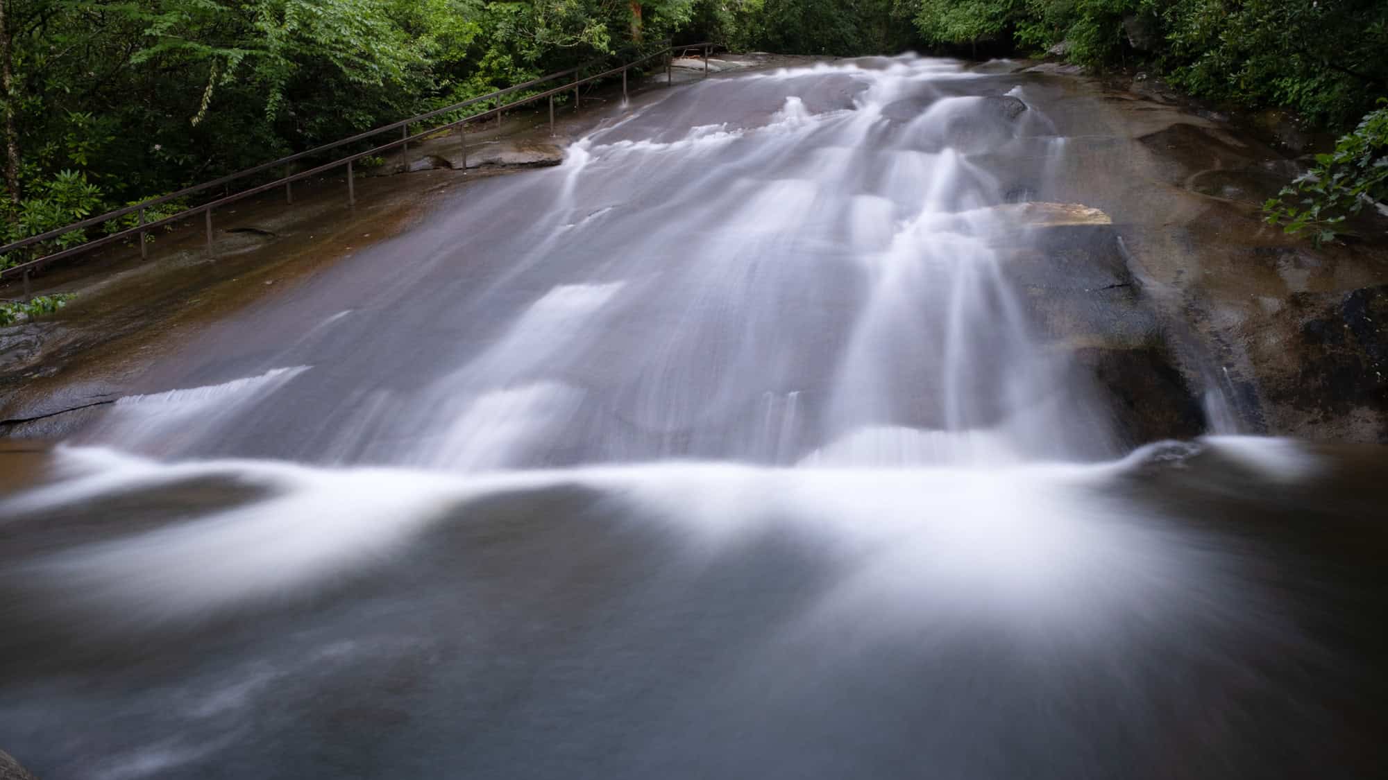 A smooth, sloped rock formation where water glides down in a natural waterslide, flanked by a wooden railing and lush green trees.