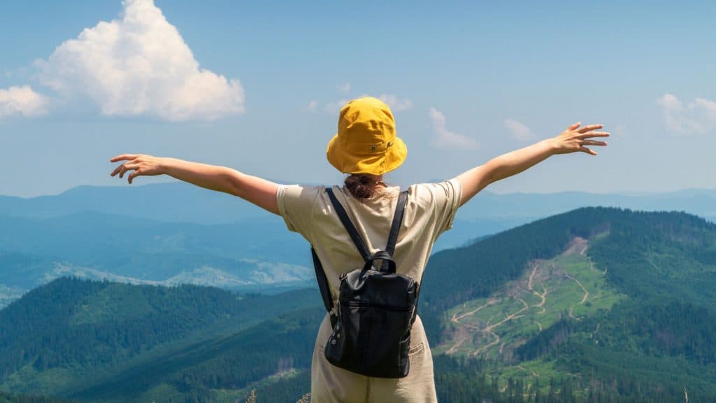 A solo traveler with a yellow bucket hat stands facing away from camera in front of a hilly green landcape.
