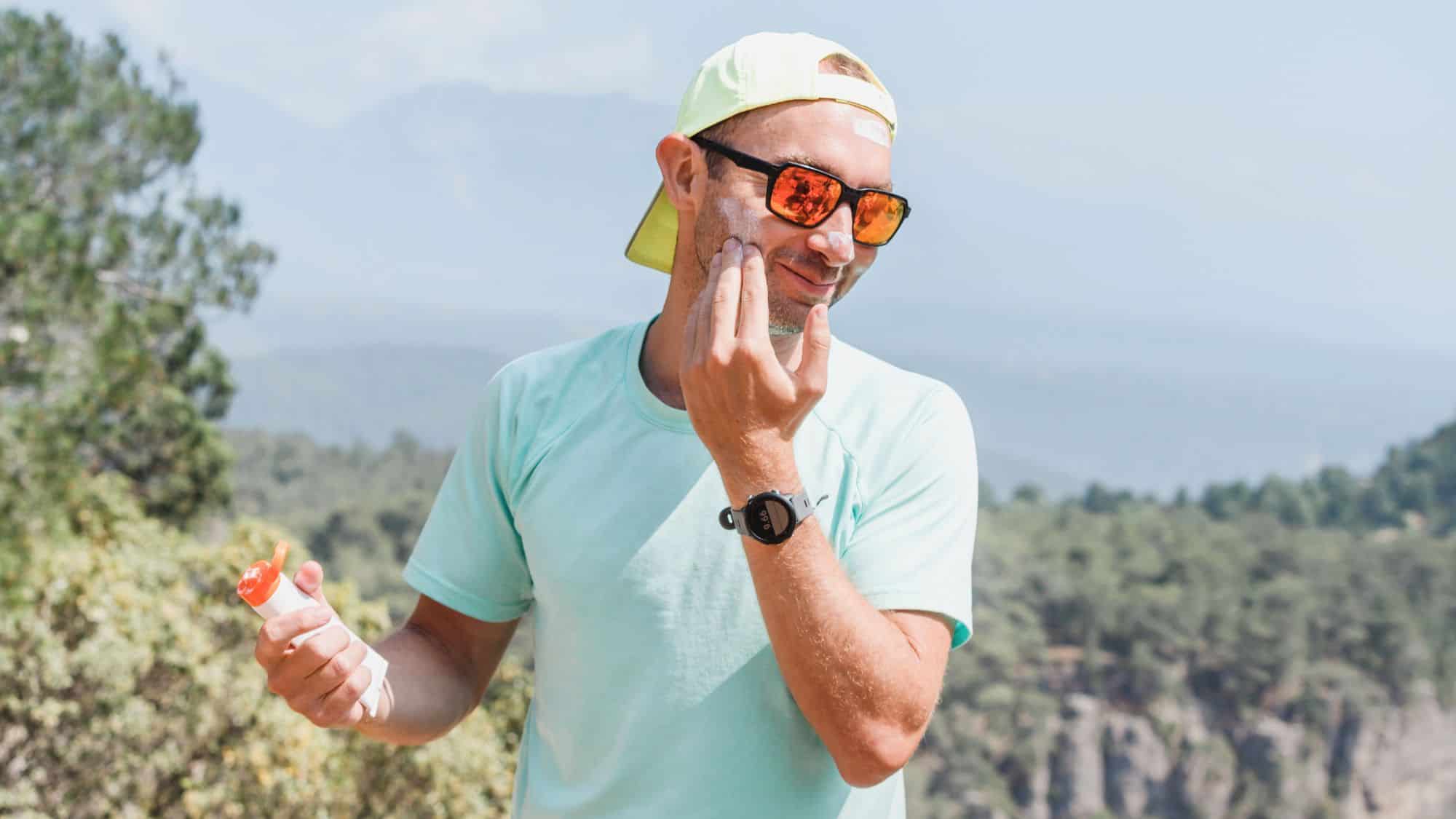 Man in a light blue shirt and cap applying sunscreen to his face outdoors, with a forested mountain range visible behind him.