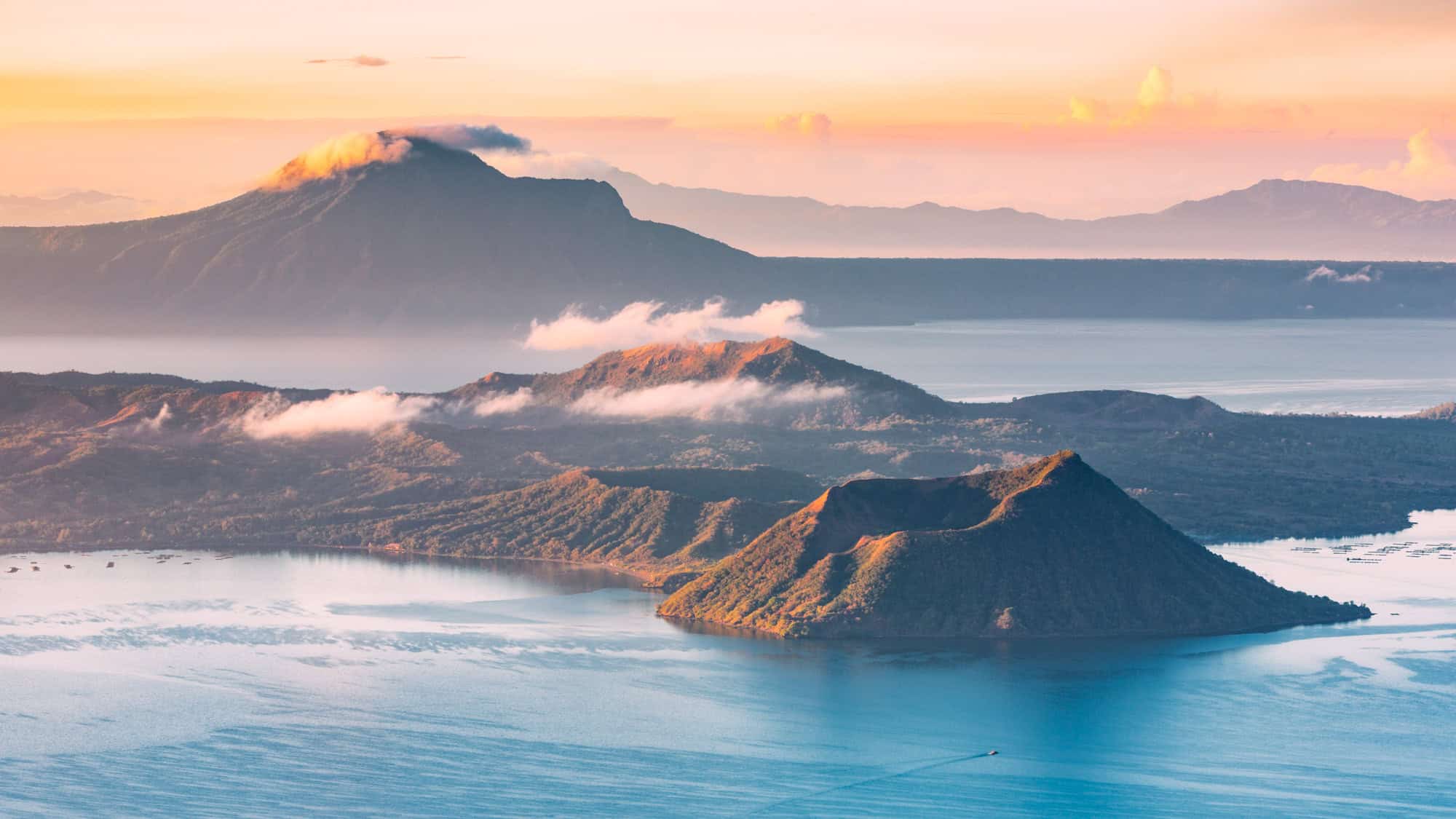 A picturesque aerial scene of the Taal Volcano in the Philippines, where a crater lake surrounds a volcanic island, backed by a larger mountain, bathed in warm sunrise hues.