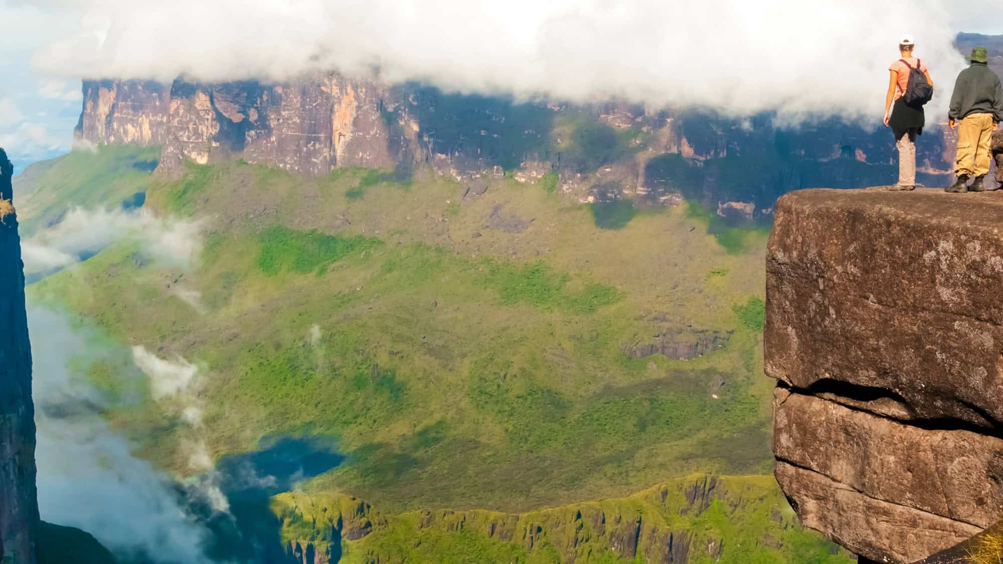 Two hikers stand on the edge of a dramatic cliff overlooking Venezuela’s mist-shrouded tepui mountains, with vertical rock walls and lush valleys below.