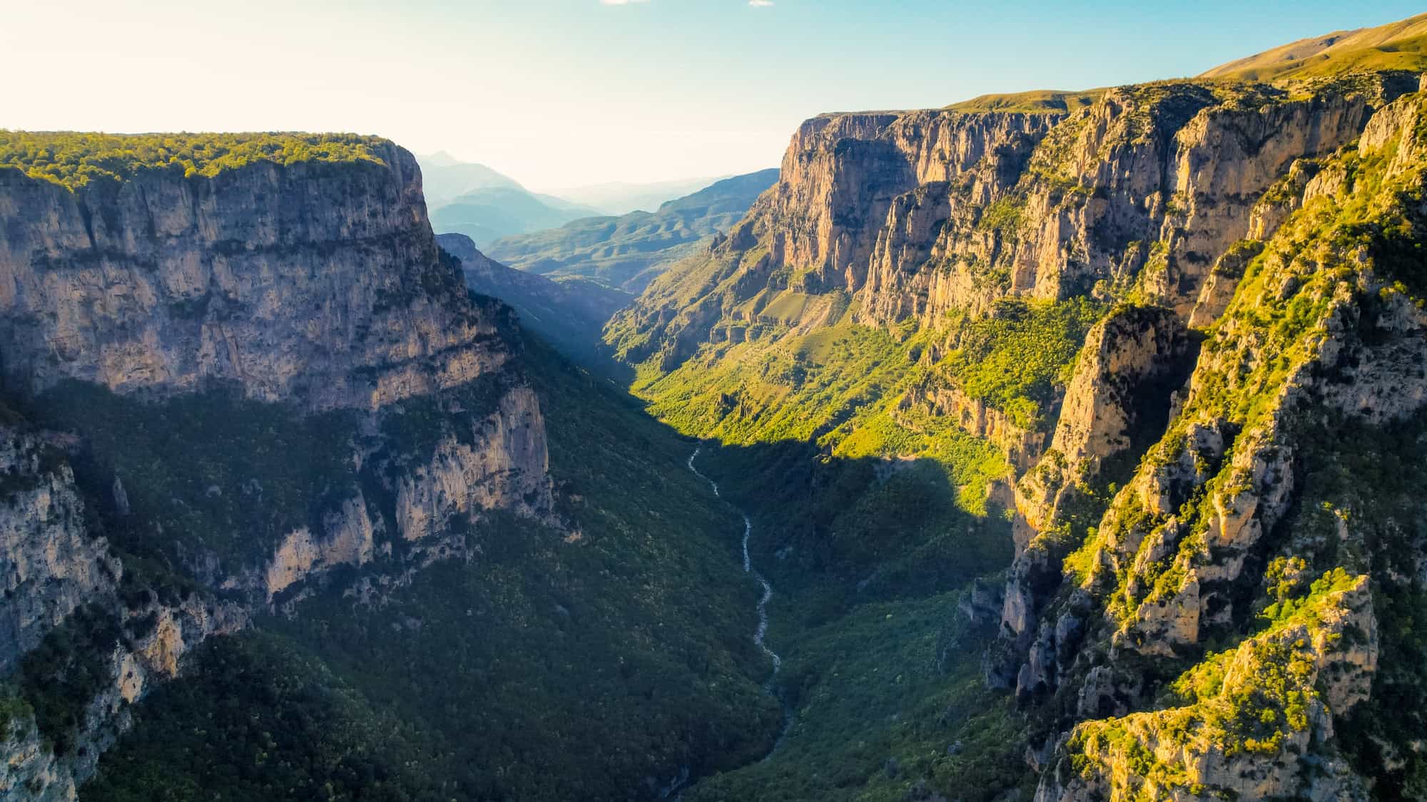 An aerial view of the lush, steep cliffs of Vikos Gorge in Greece, with dramatic shadows cast over the deep valley and winding river below.


