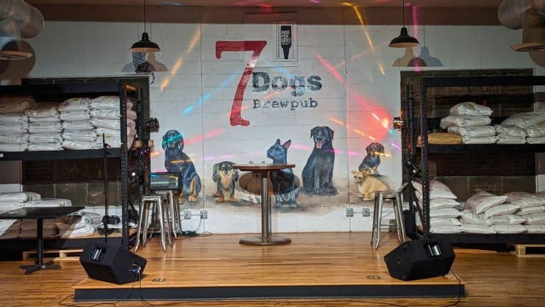 A cozy stage setup inside 7 Dogs Brewpub features a painted mural of various dog breeds on a white brick wall, with sacks of brewing ingredients stacked on shelves. Disco lights reflect gently across the room, adding color to the casual brewery scene.