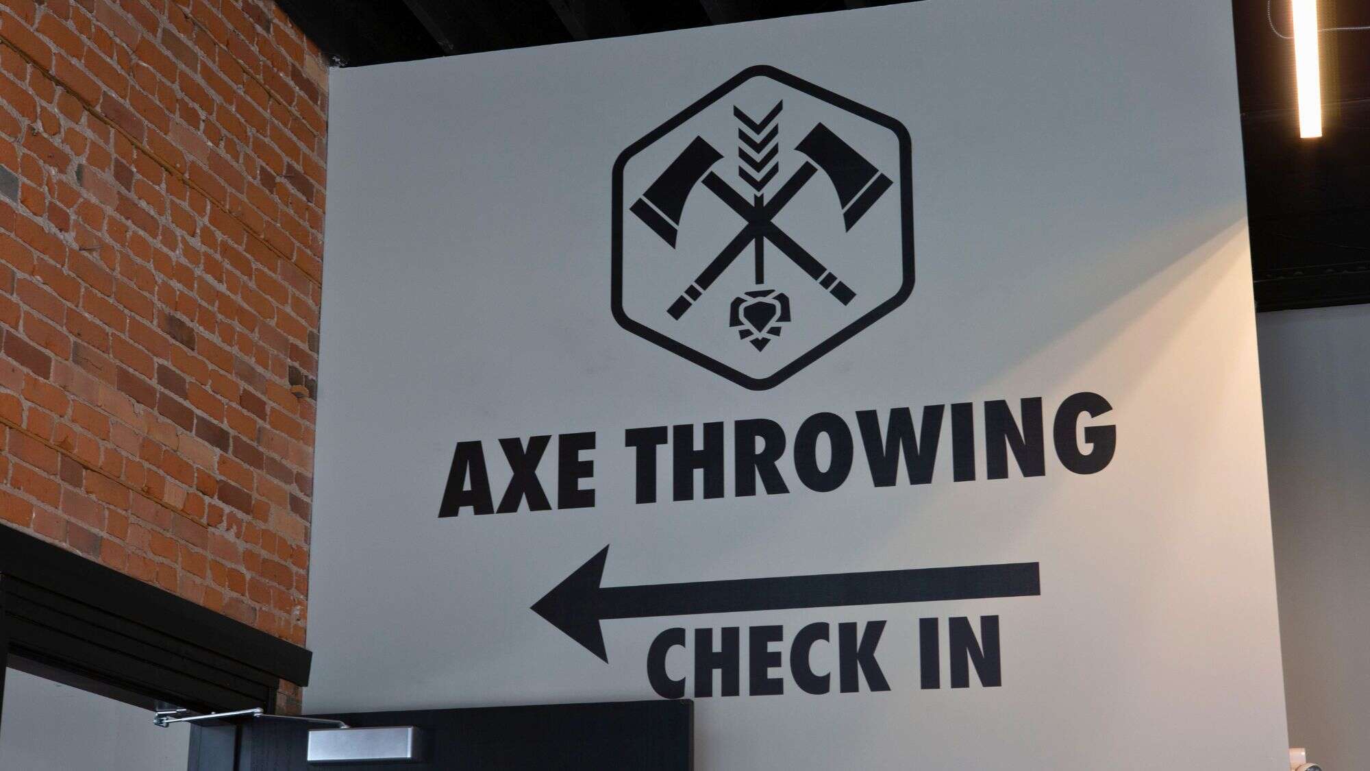 Indoor wall signage directing guests to check in for an axe throwing experience, featuring a black-and-white design with crossed axes and a bold arrow pointing left.