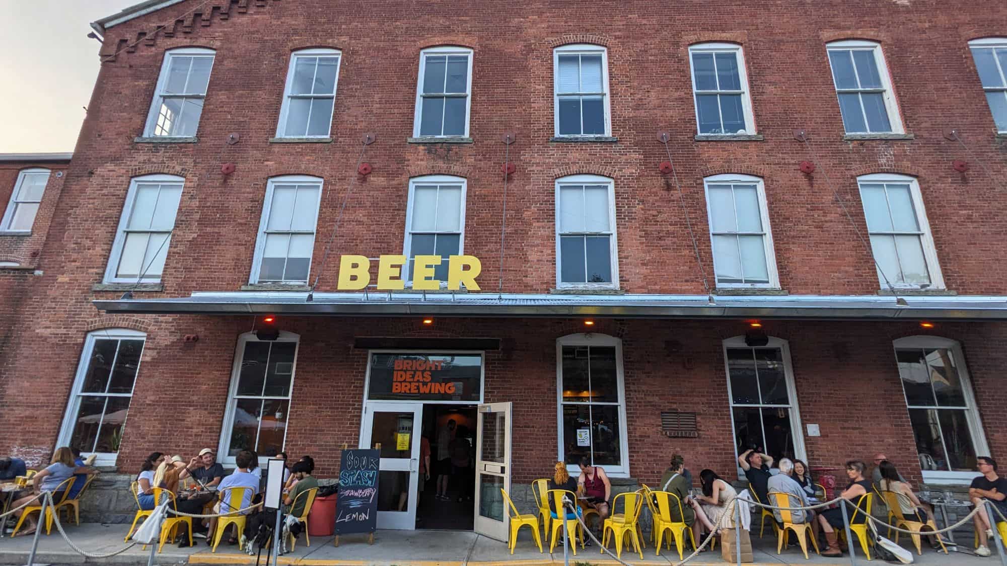 Front of a red-brick building with a large yellow "BEER" sign above the entrance to Bright Ideas Brewing, where patrons are seated at yellow chairs enjoying drinks outdoors.