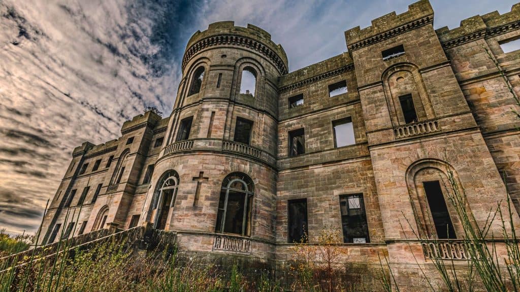A moody, low-angle view of the ruined Dalquharran Castle in Scotland, with its weathered stone facade, empty window frames, and a turret rising dramatically under a textured sky.