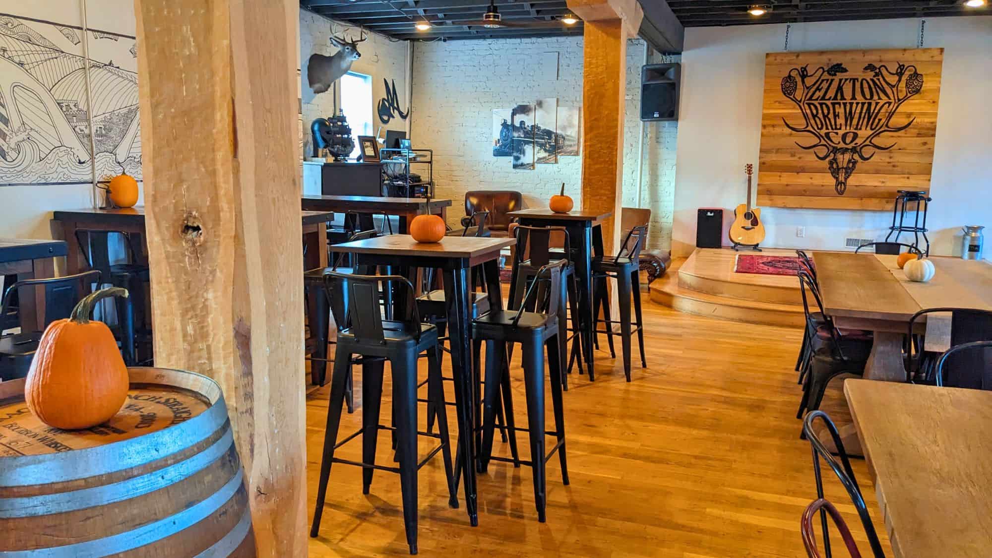 Cozy taproom with warm wooden floors, high tables topped with pumpkins, and a small performance stage with a guitar, set against a wall bearing the Elkton Brewing Co. logo.