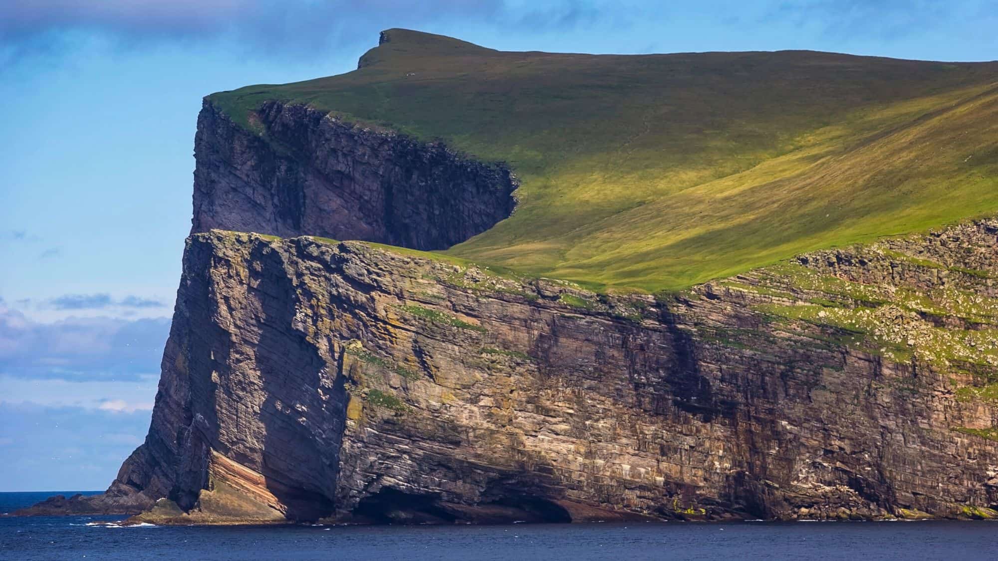 A dramatic cliffside view of Foula Island in Shetland, Scotland, with steep rock faces rising sharply from the North Atlantic and a grassy, gently sloping plateau above.