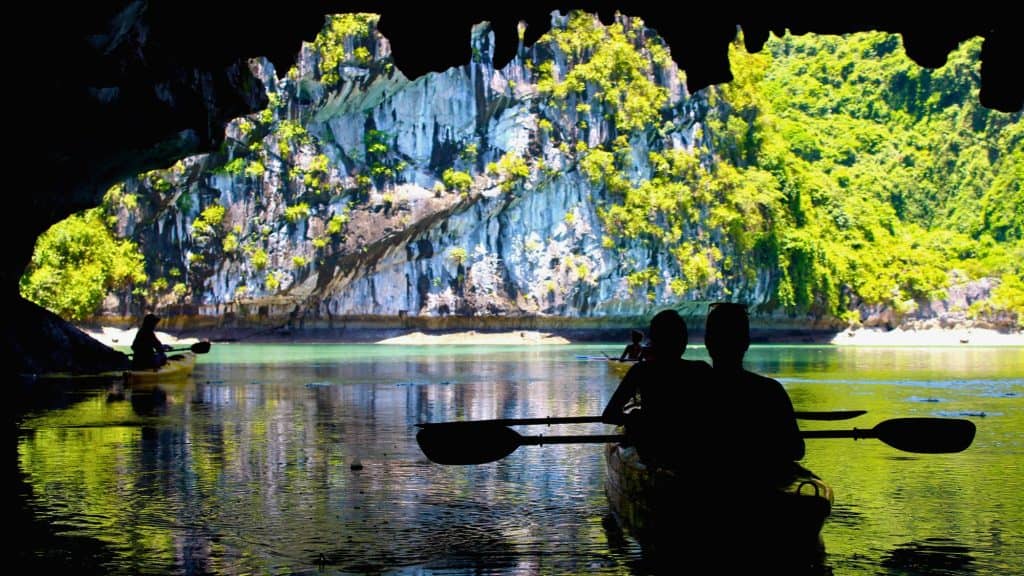 Silhouetted kayakers paddle through a dark cave into the sunlight, revealing towering limestone cliffs covered in lush greenery reflecting on calm turquoise waters.