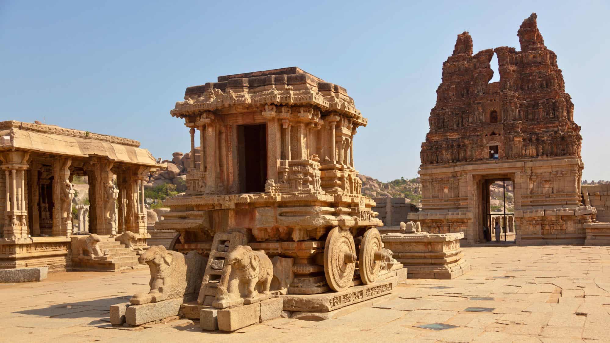 A sandstone chariot carved in great detail sits in the courtyard of the Vittala Temple complex in Hampi, India, flanked by majestic ruined temple structures under a clear sky.