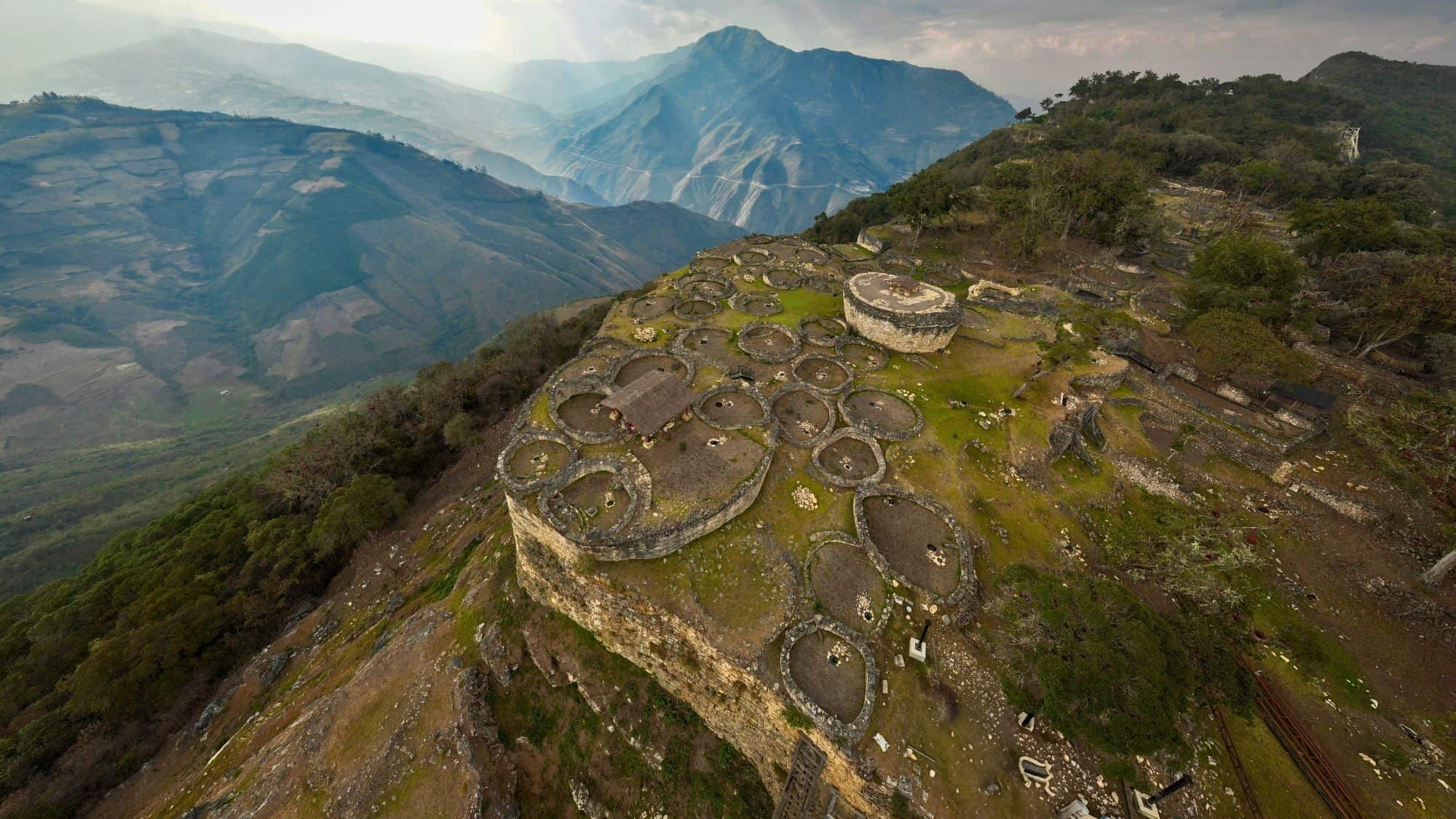 Circular stone foundations of an ancient settlement sprawl across a mountaintop at the Kuelap Fortress in Peru, with dramatic views of forested valleys and distant peaks.