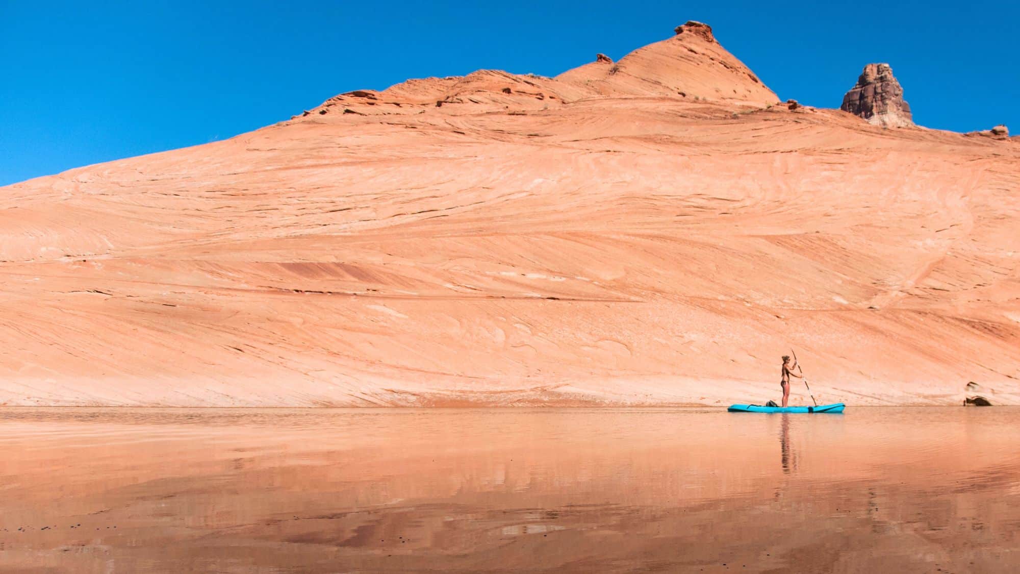 A woman stands on a bright blue paddleboard at the edge of Lake Powell, dwarfed by the sweeping curves of rust-red sandstone cliffs under a clear blue sky.