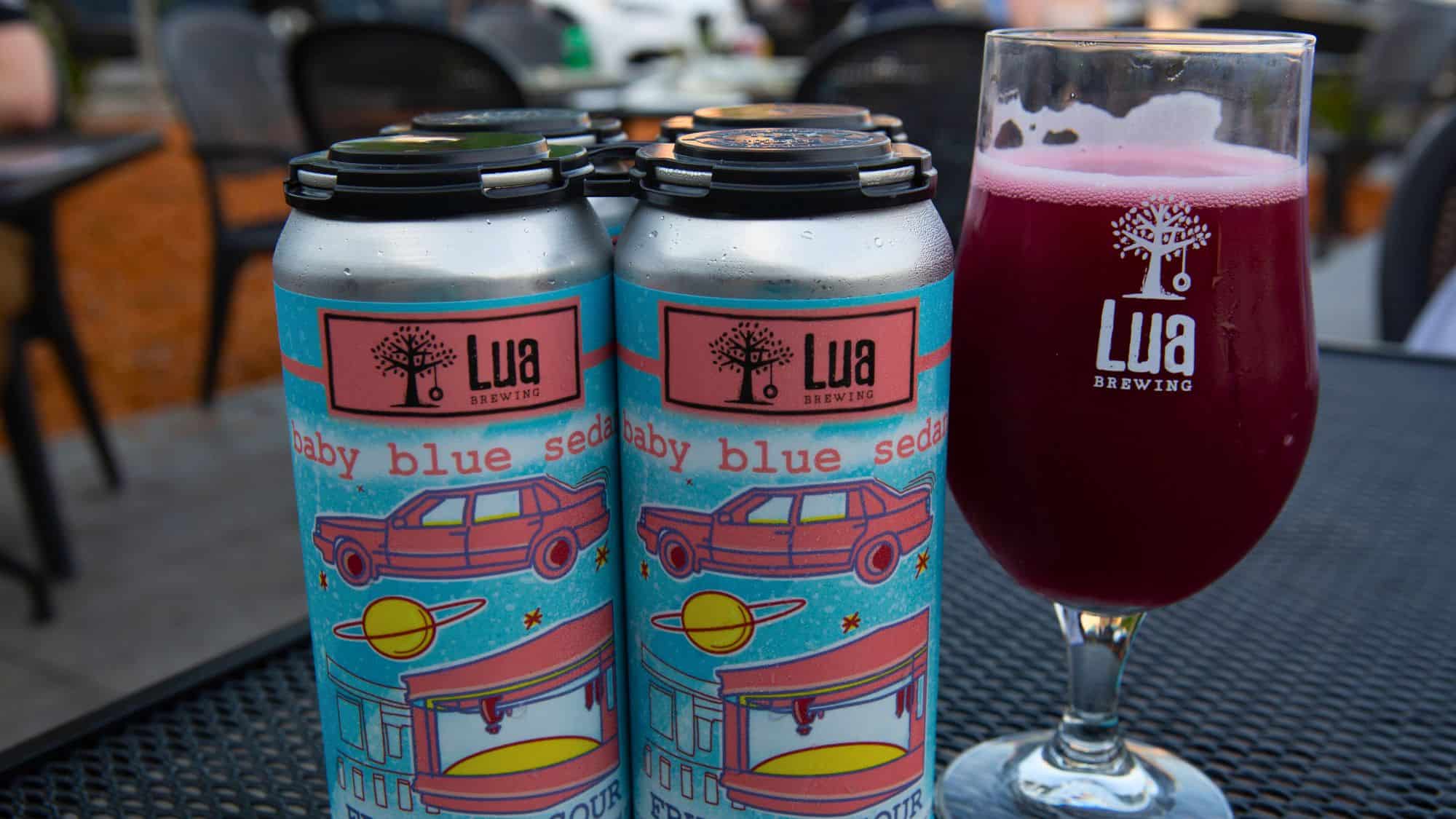Close-up of Lua Brewing's "Baby Blue Sedan" sour beer, showing a vibrant can design with a red car and planet illustrations next to a glass of red-hued beer with the Lua logo.