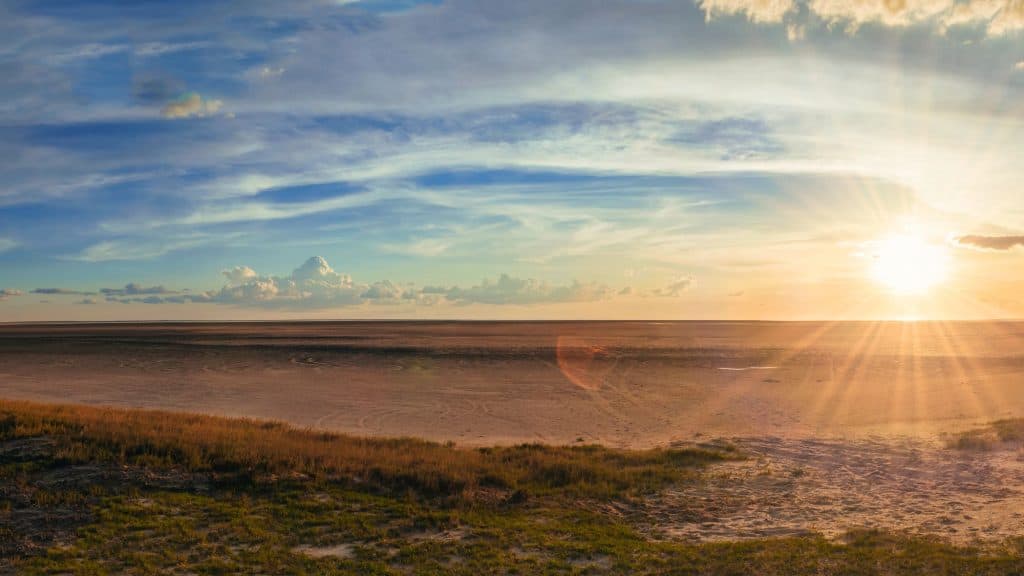 A vast, flat salt pan stretches beneath a wide open sky at sunset in the Makgadikgadi Pans of Botswana, with lens flare from the sun casting warm rays over the cracked earth and grasses.