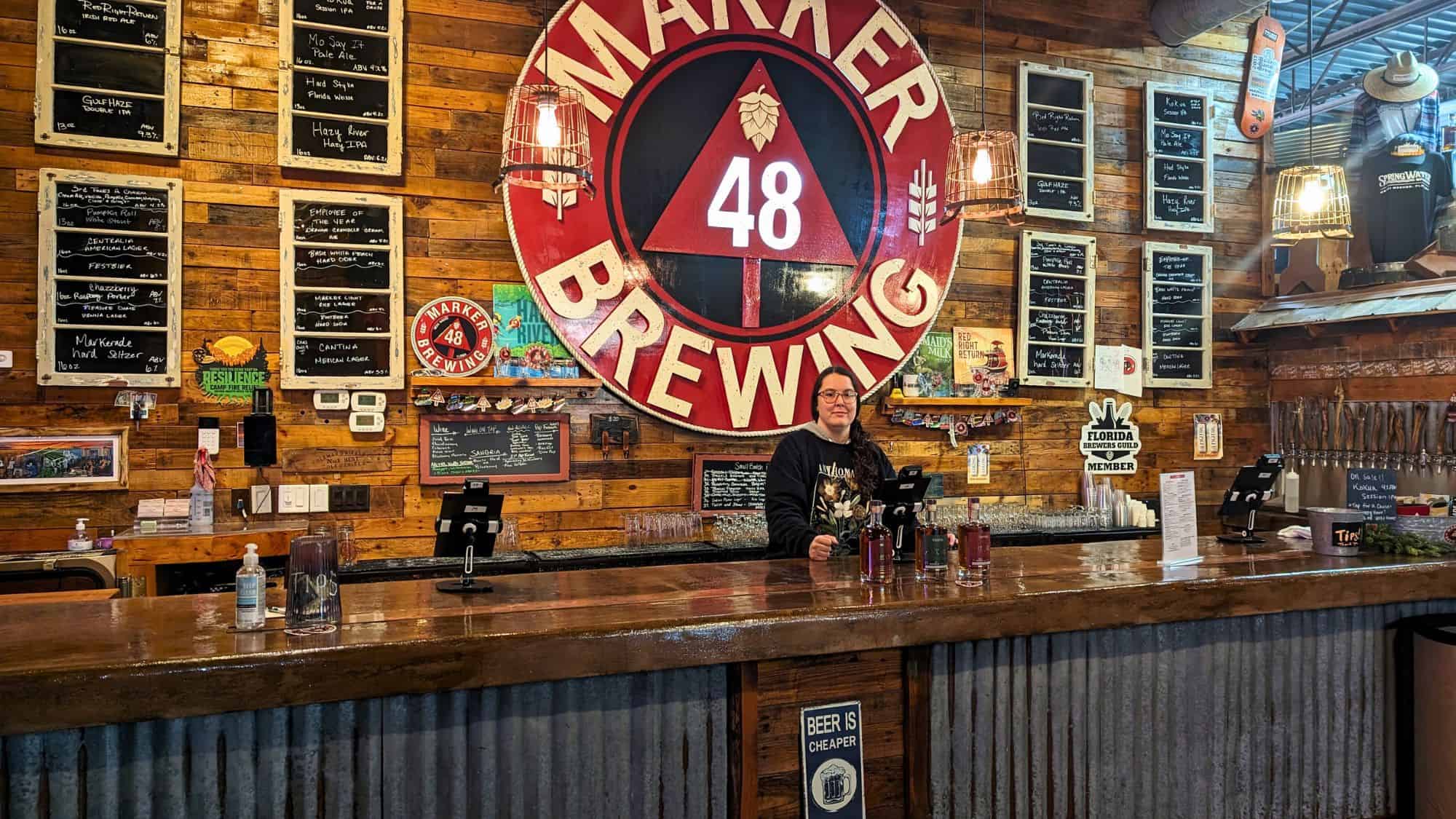 Rustic wood-accented taproom with a large red Marker 48 Brewing sign, chalkboard beer menus, and a bartender standing behind a glossy wood bar.