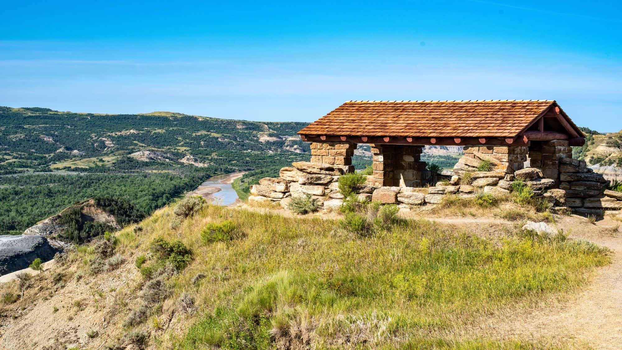 A rustic stone shelter with a wooden roof overlooks a winding river and expansive green valley below, set against a backdrop of layered cliffs.