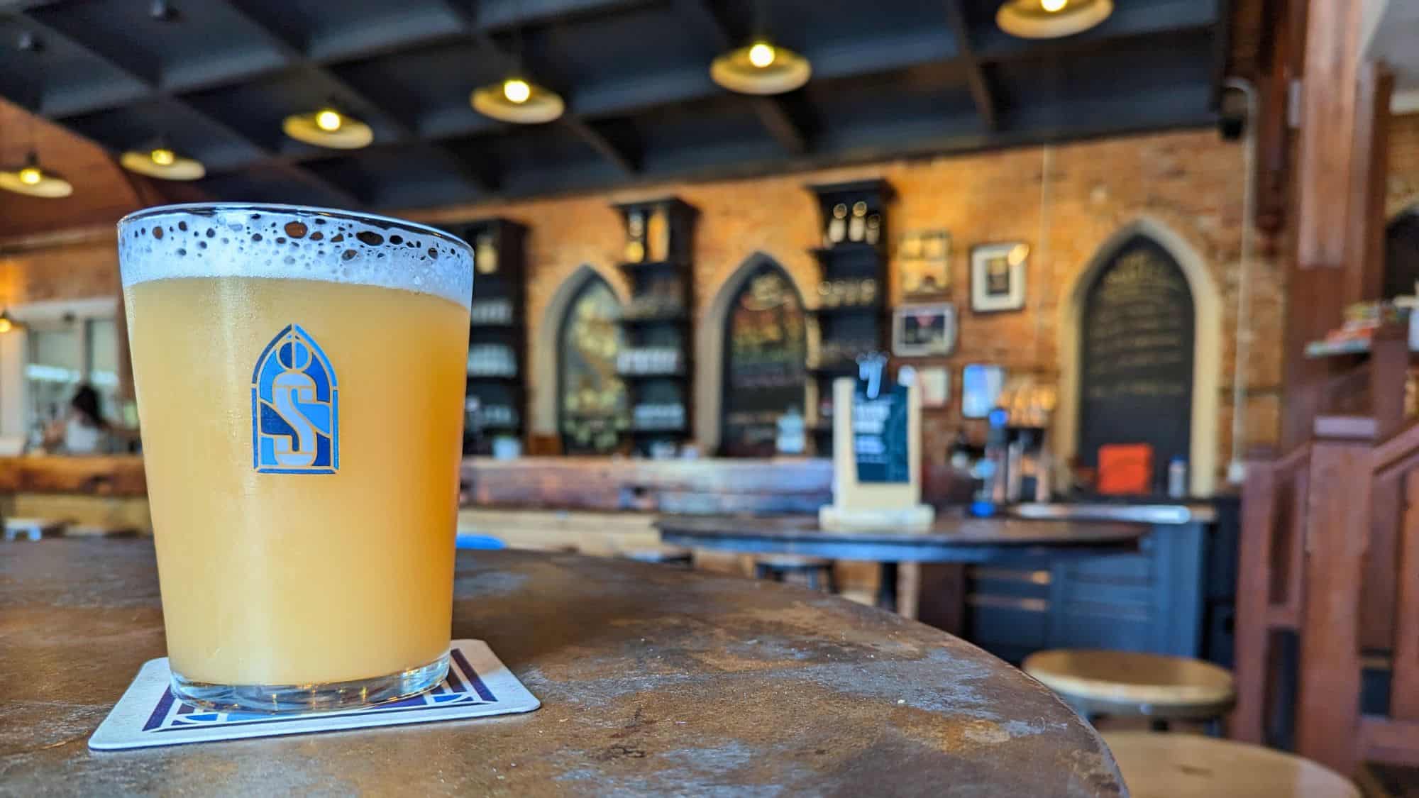 Close-up of a hazy golden beer in a branded glass from Silversmith Brew, set on a coaster in a cozy bar with gothic-style arched shelves in the background.