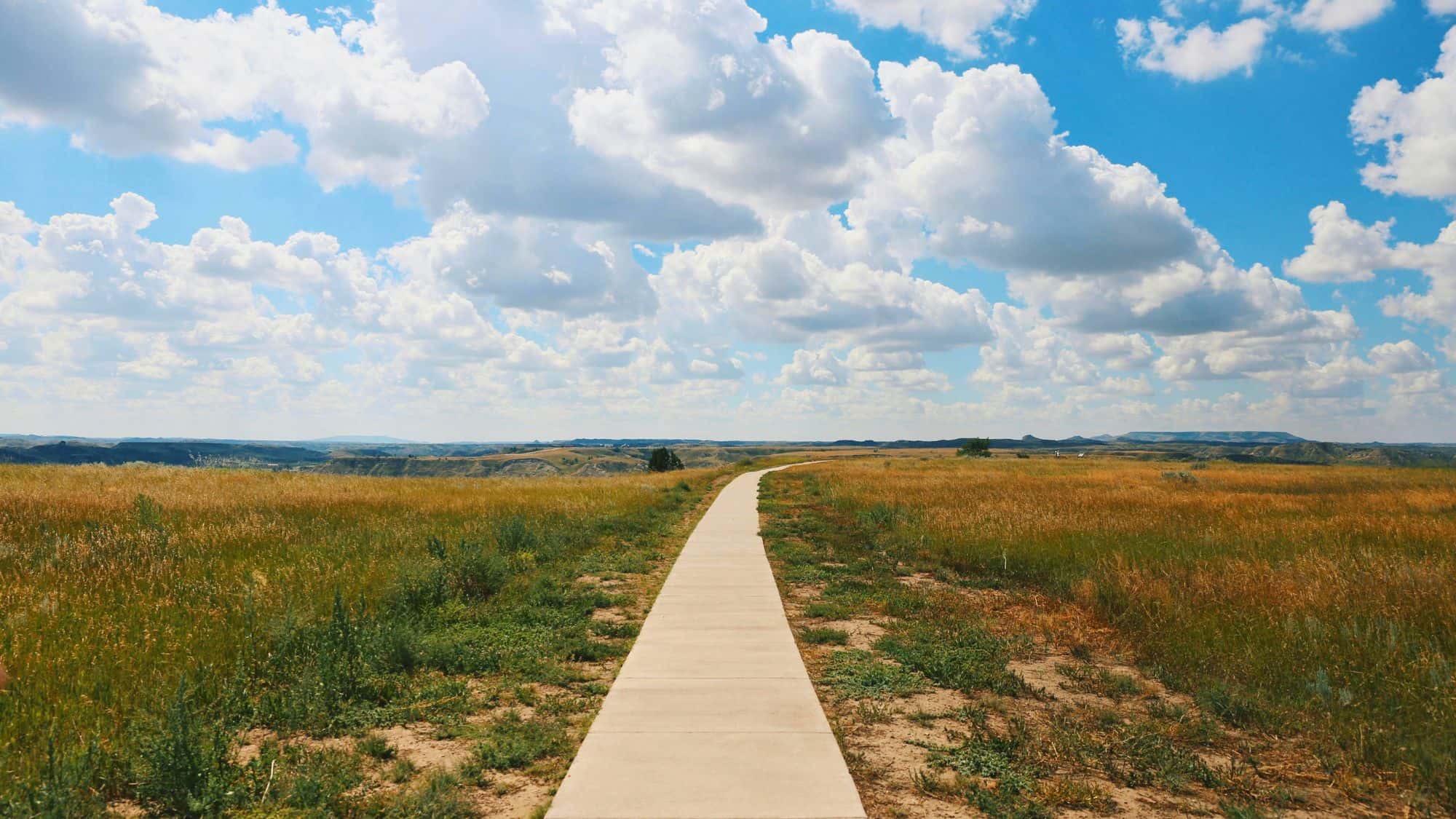 A paved walking path cuts through grassy plains under a sky filled with puffy clouds, leading toward the rolling hills in the distance.