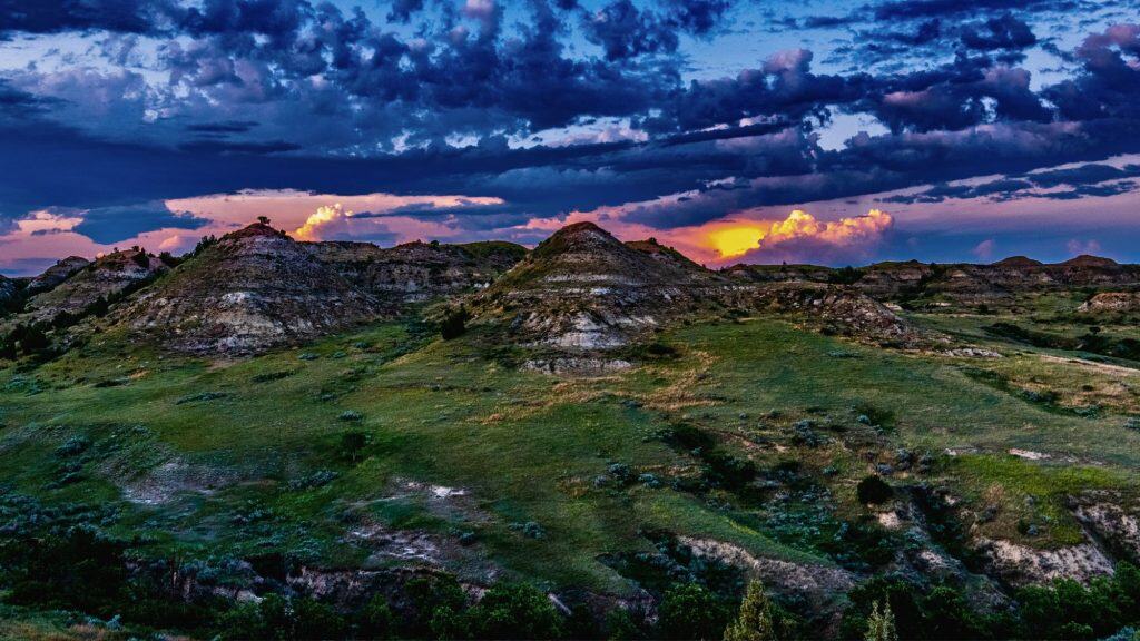 A dramatic sunset bathes the rugged badlands in rich purples and oranges, with cloud-covered peaks glowing against the fading daylight.