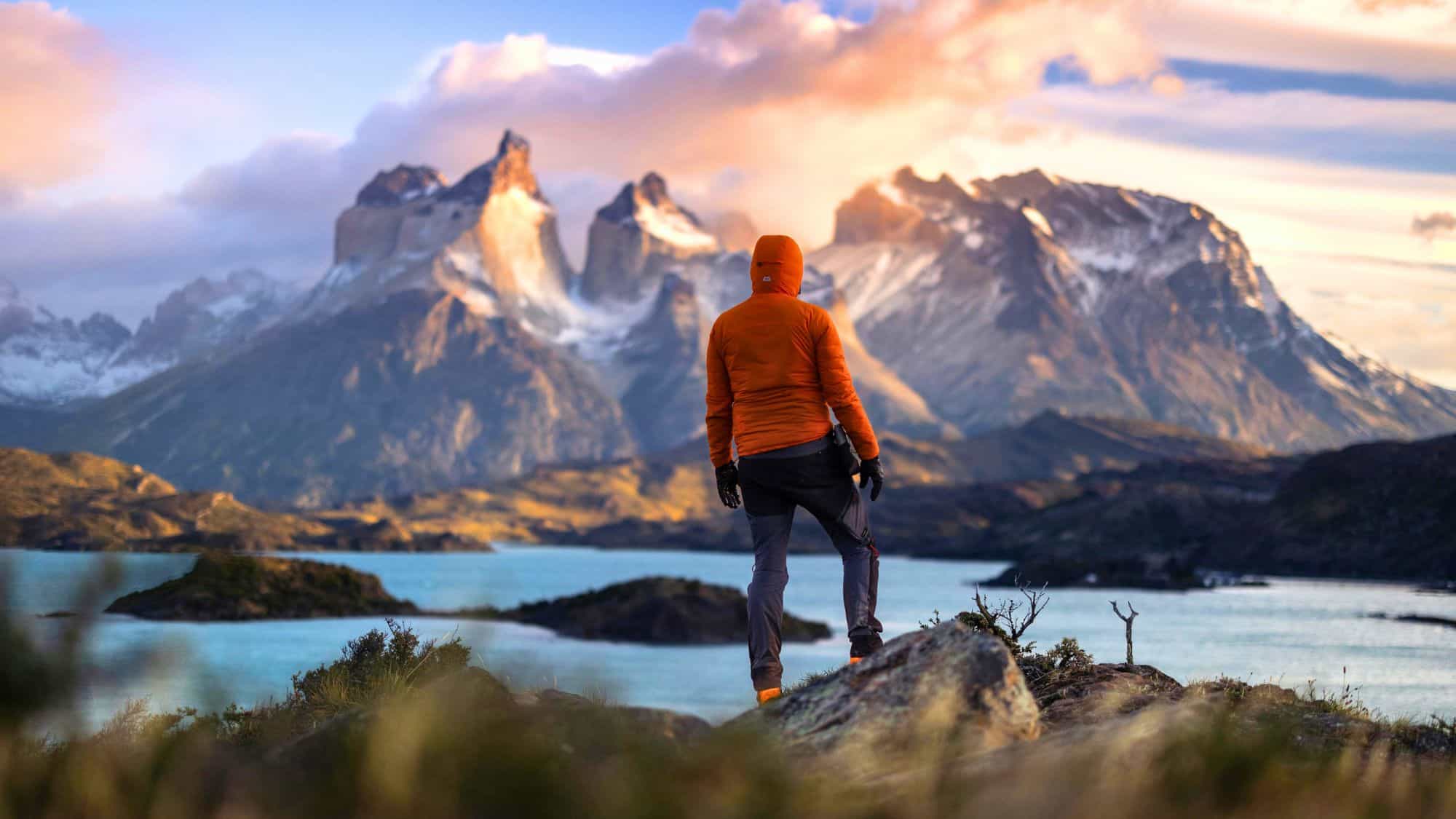 A hiker in an orange jacket stands on a rocky hilltop at dawn, gazing across a scenic expanse of icy blue lakes and the dramatic, craggy peaks of Torres del Paine lit by warm sunrise hues.