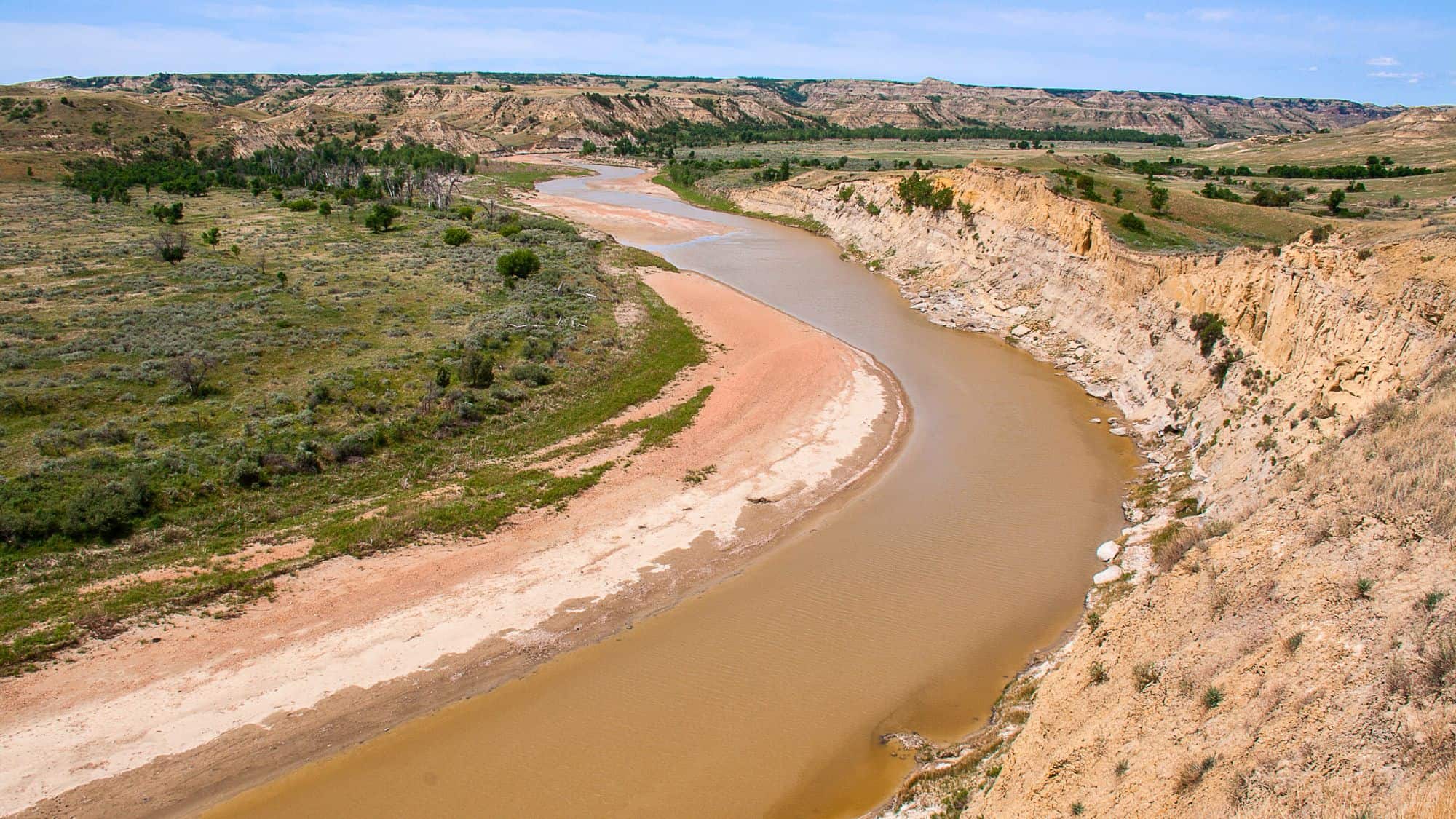 A gently curving river winds through a canyon of beige cliffs and green sagebrush, carving through the North Dakota landscape under a soft blue sky.
