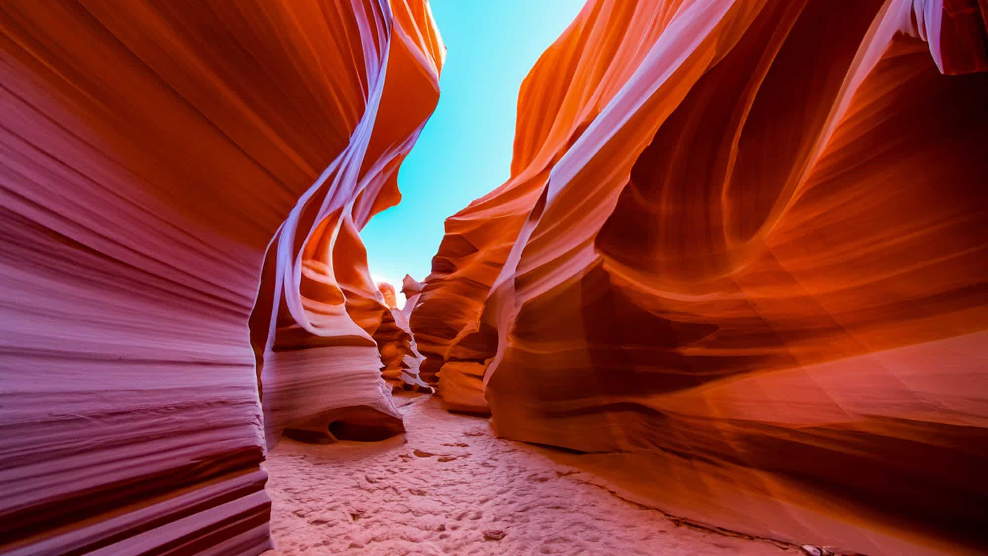 A surreal corridor of Antelope Canyon, with vibrant swirls of orange and pink sandstone carved smooth by wind and water. Bright blue sky peeks through the tight curves above.