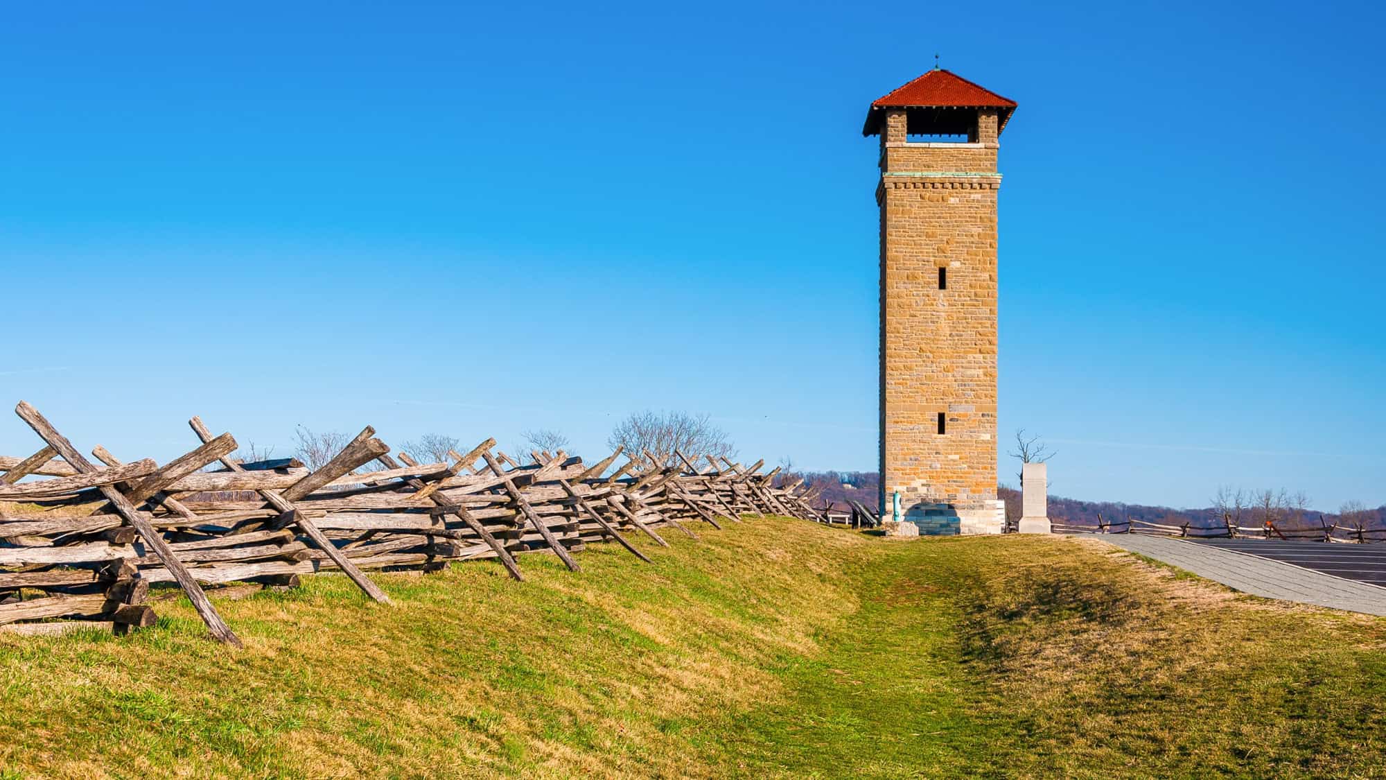 A tall stone observation tower with a red roof stands near a zigzag split-rail fence at Antietam National Battlefield, under a vivid blue sky.