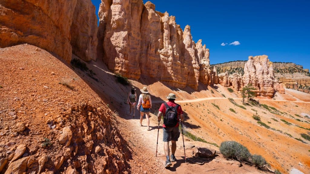 Three hikers trek along a winding trail beneath massive orange and pink hoodoos in Bryce Canyon. The vivid rock formations and deep blue sky create a dramatic Southwestern desert scene.