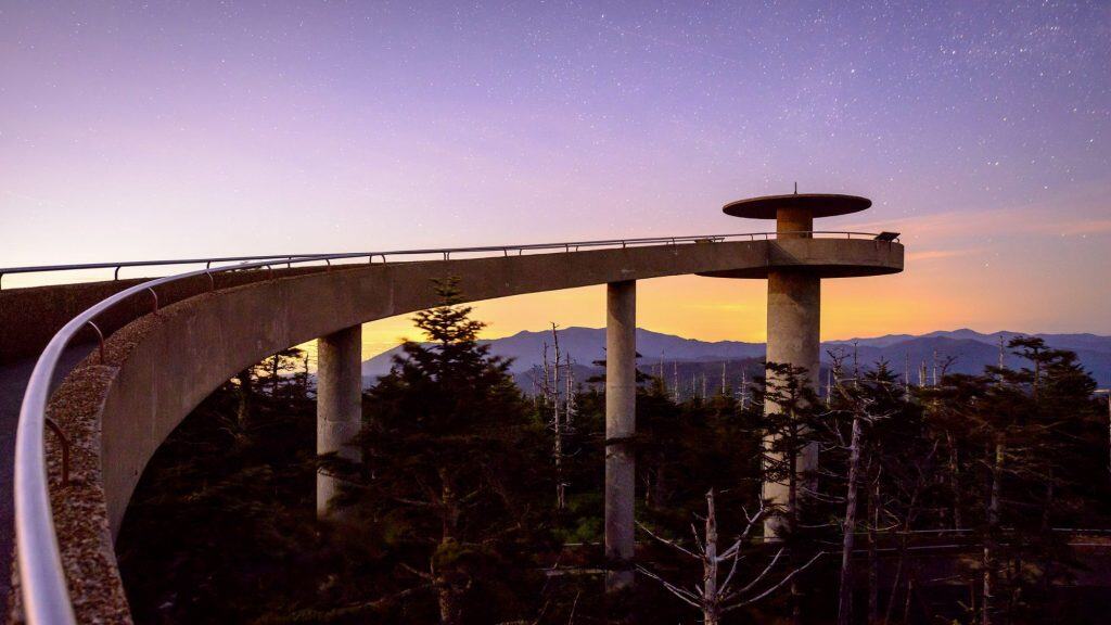 The Clingmans Dome tower is bathed in golden and purple twilight hues, with stars faintly visible above and the ramp gently curving through silhouetted treetops and mountain ridges beyond.