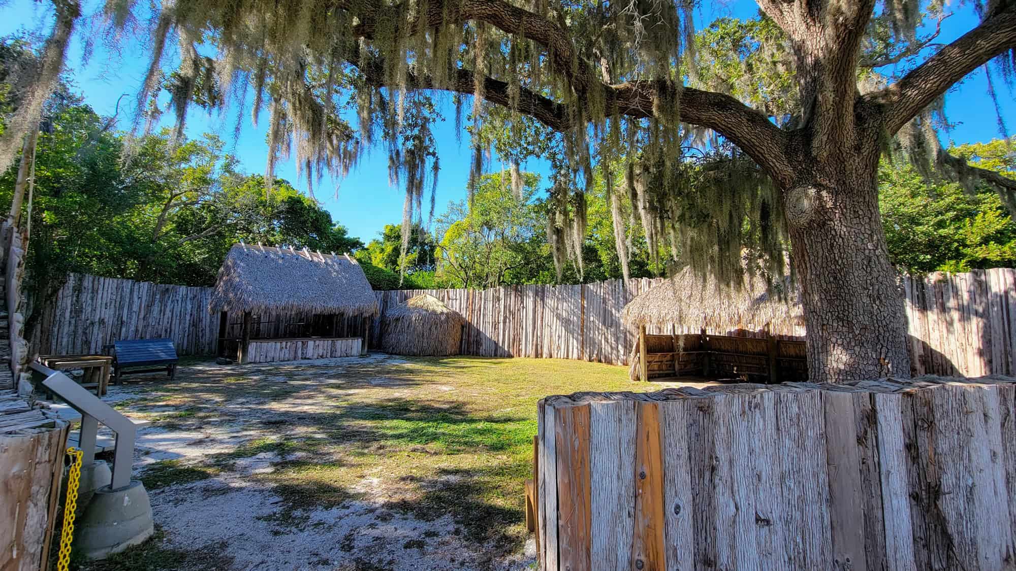 A quiet clearing under a large tree draped in Spanish moss at De Soto National Memorial, enclosed by a wooden palisade and traditional chickee-style shelters.