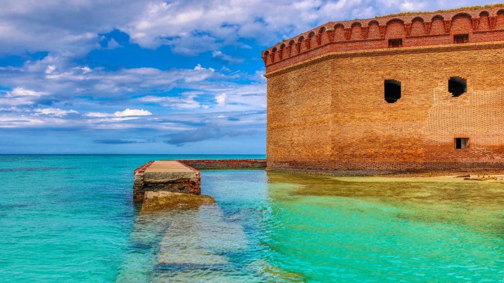 Bright turquoise water laps against the massive red-brick walls of Fort Jefferson at Dry Tortugas National Park, under a partly cloudy sky in the remote Florida Keys.