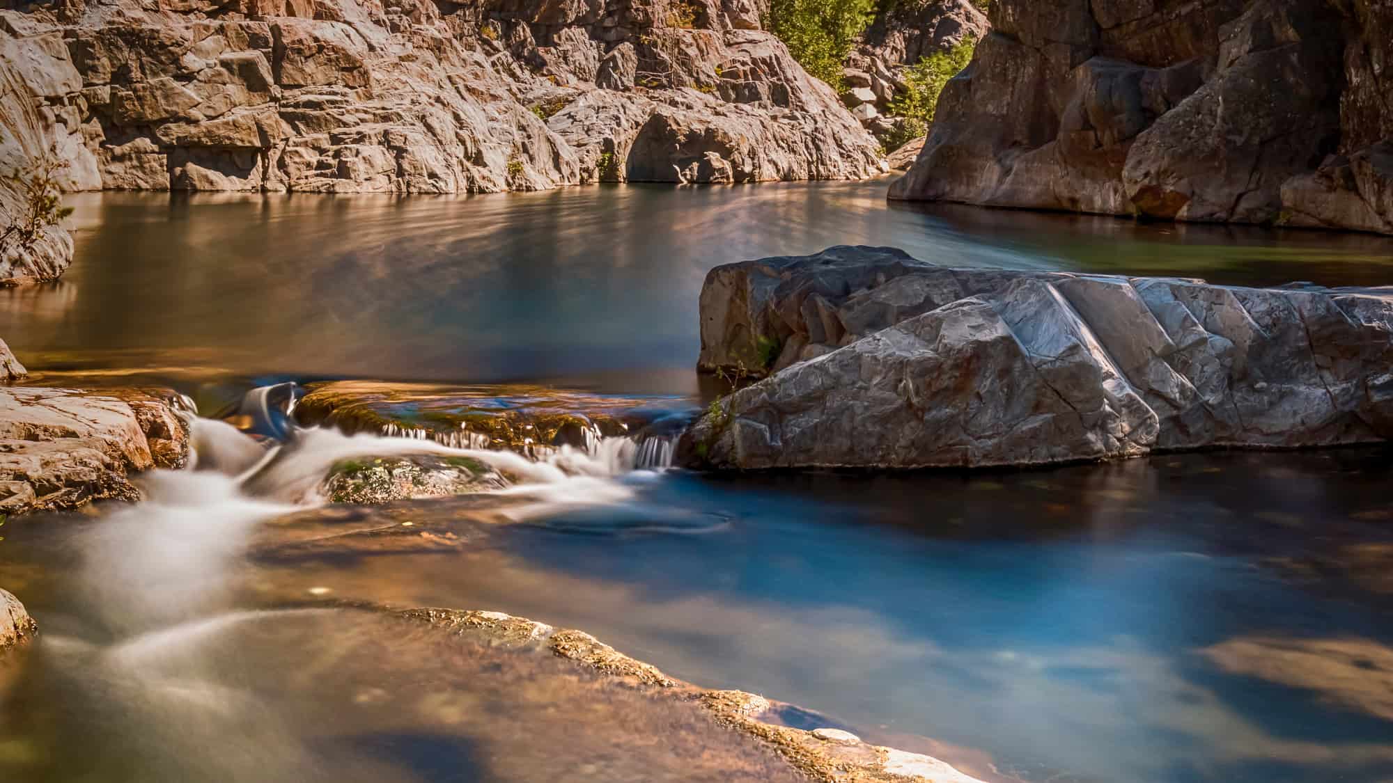 A serene pool with crystal-clear water flowing gently over smooth rocks, surrounded by steep canyon walls. The water reflects the golden sunlight and the tranquil mood.